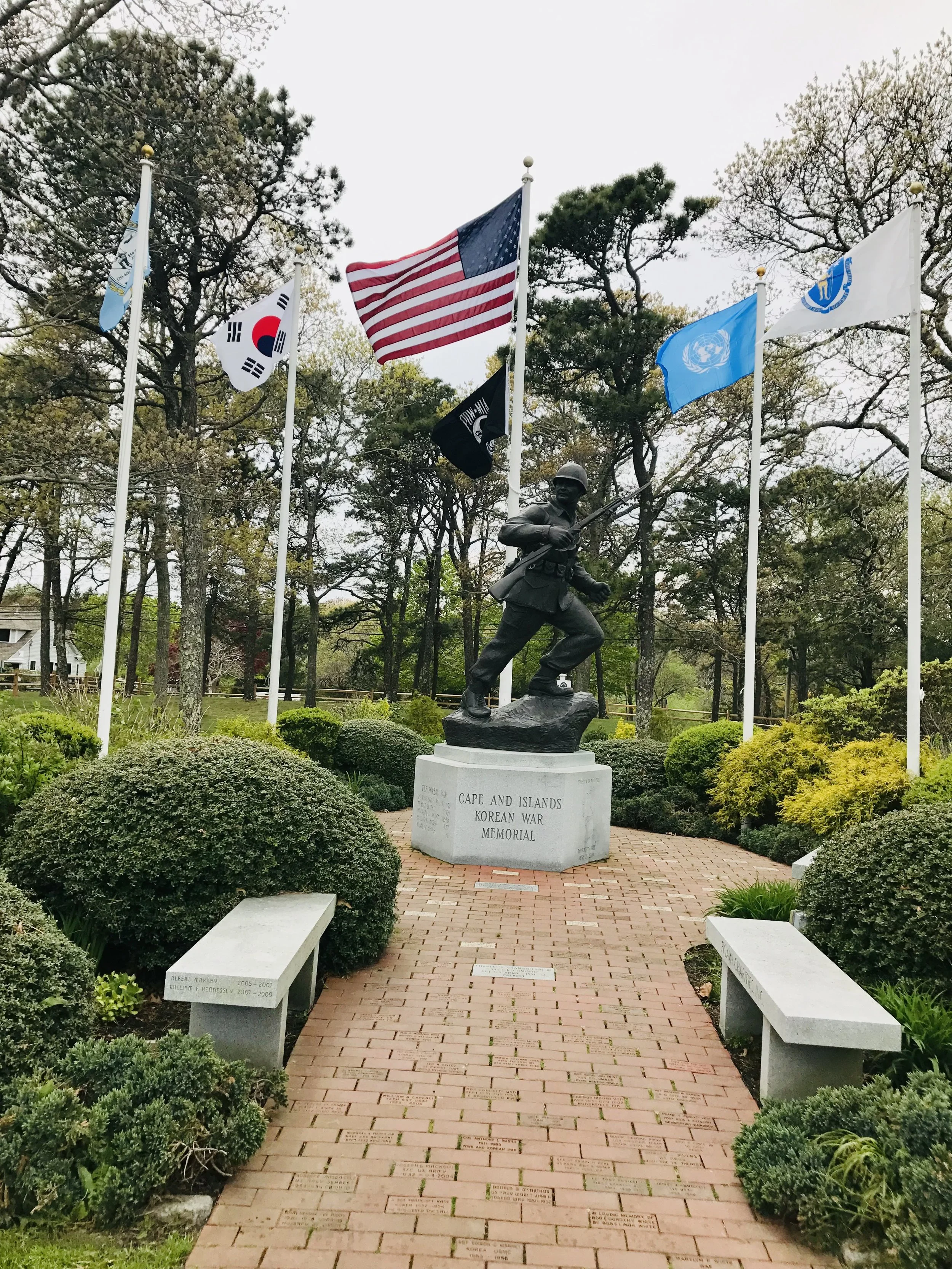 A monument at the Cape and Islands Korean War Memorial with a soldier statue, surrounded by six flags including the American flag, South Korea flag, and United Nations flag, among trees and bushes.