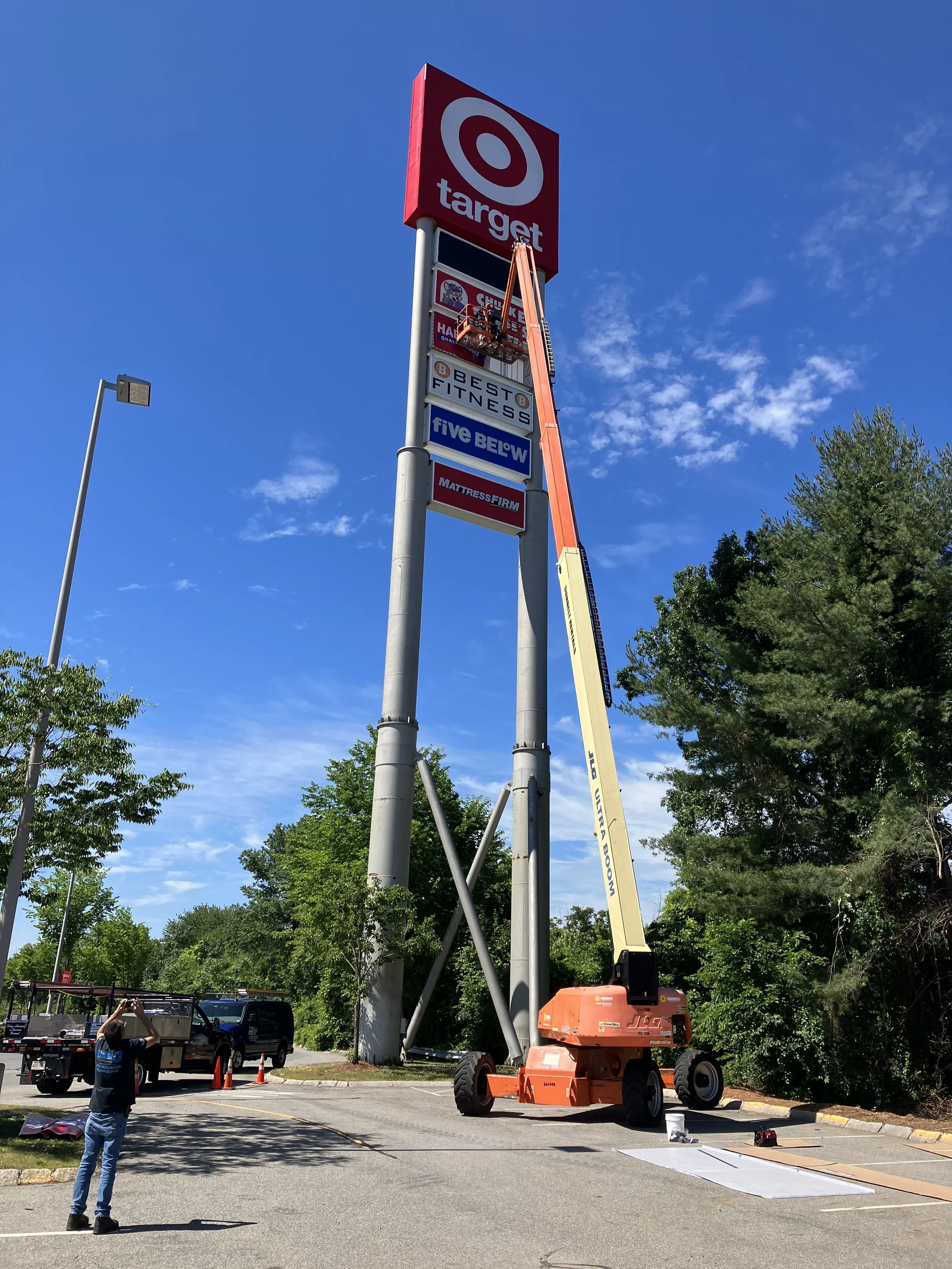 Workers installing or repairing the Target store sign on a tall pole using a cherry picker lift, with trees and parked vehicles in the background.