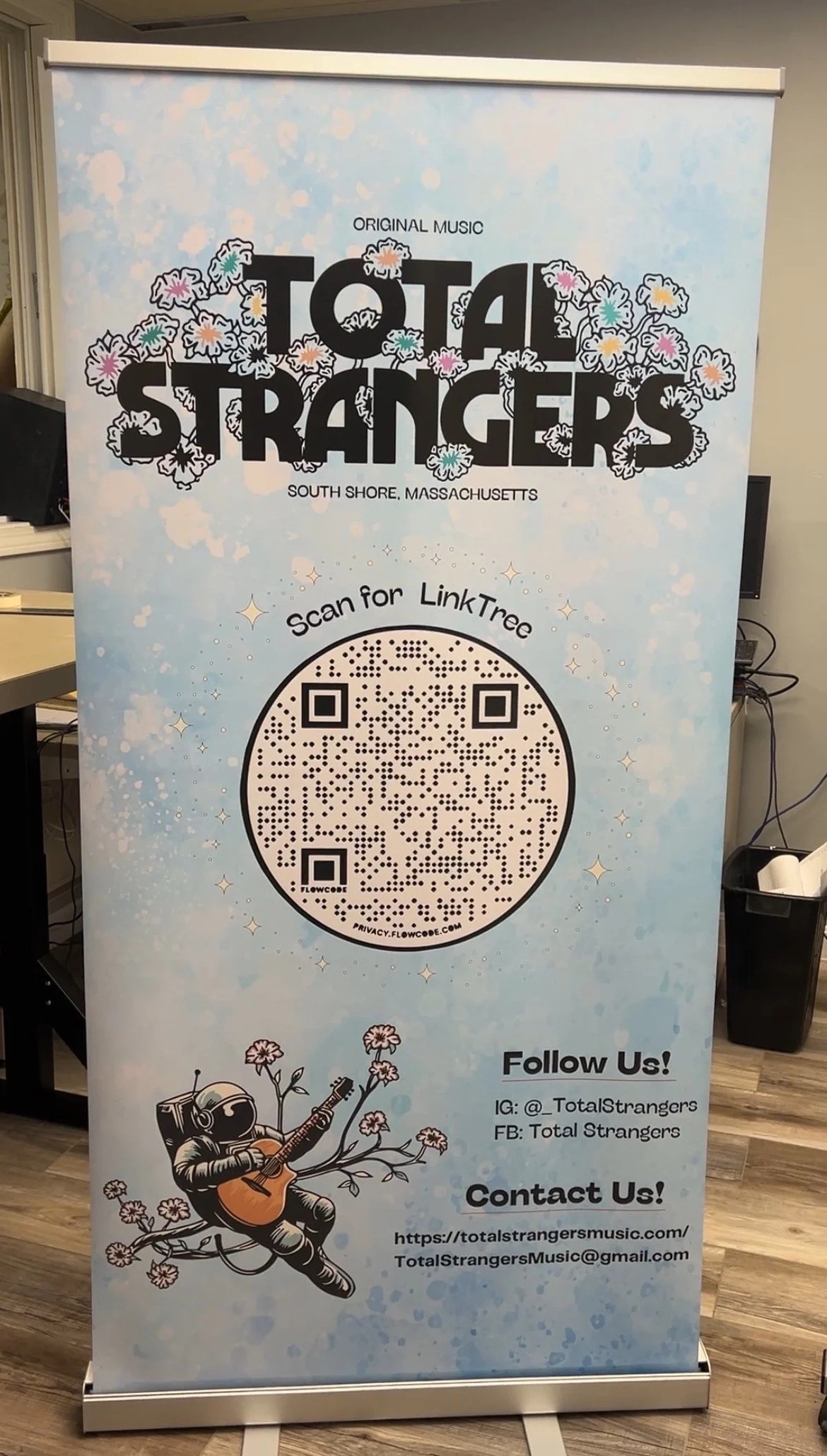Light blue banner with black text and illustrations advertising 'Total Strangers' music, based in South Shore, Massachusetts. Features a QR code with 'Scan for LinkTree,' a cartoon astronaut playing a guitar surrounded by flowers, and social media and contact information.