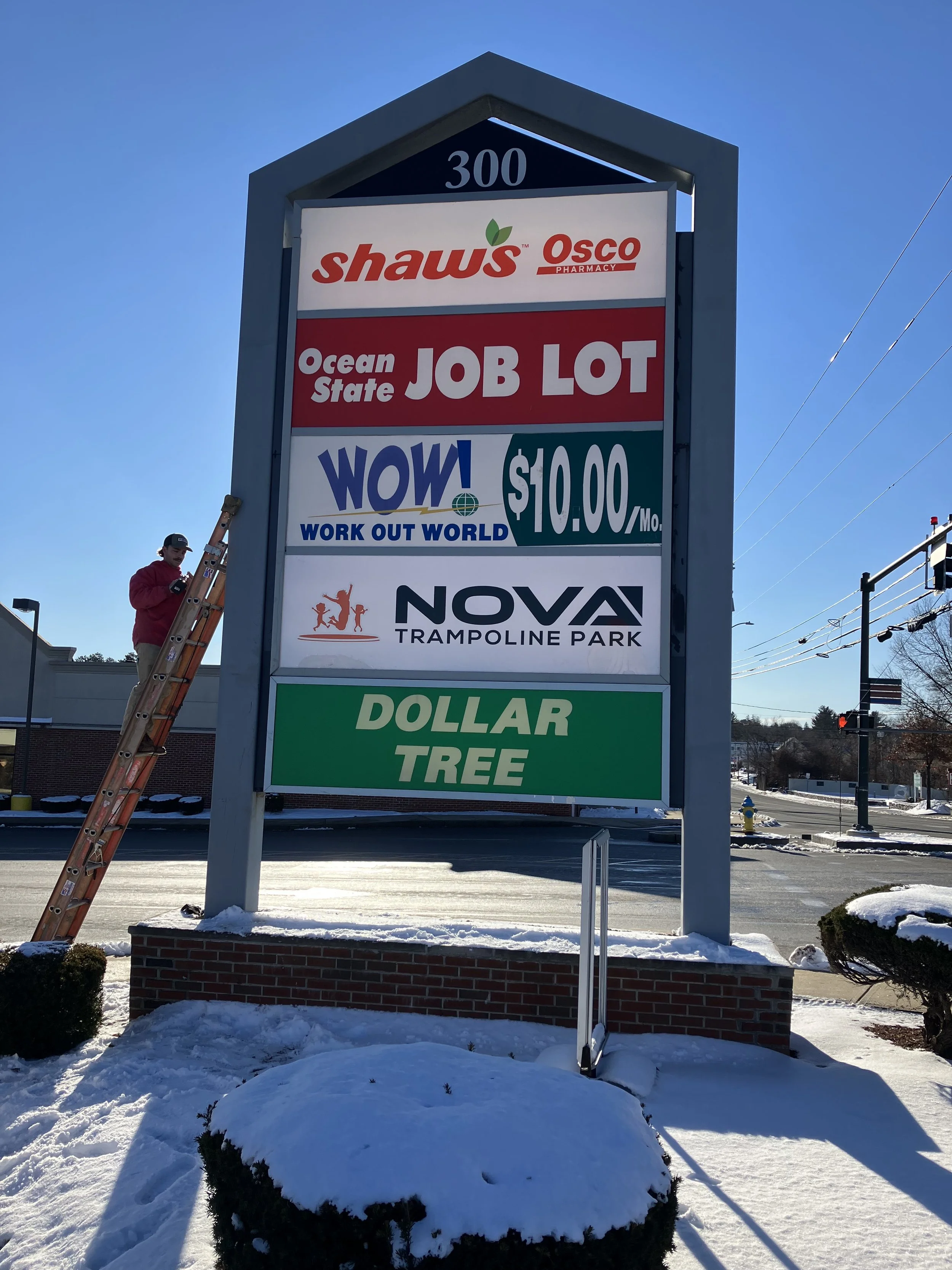 A large signboard advertising various businesses, including Shaw's Osco Pharmacy, Ocean State Job Lot, WOW Work Out World for $10.00 per month, Nova Trampoline Park, and Dollar Tree. There is a person on a ladder wearing a red jacket repairing or installing part of the sign. The ground is covered with snow and the sky is clear and blue.