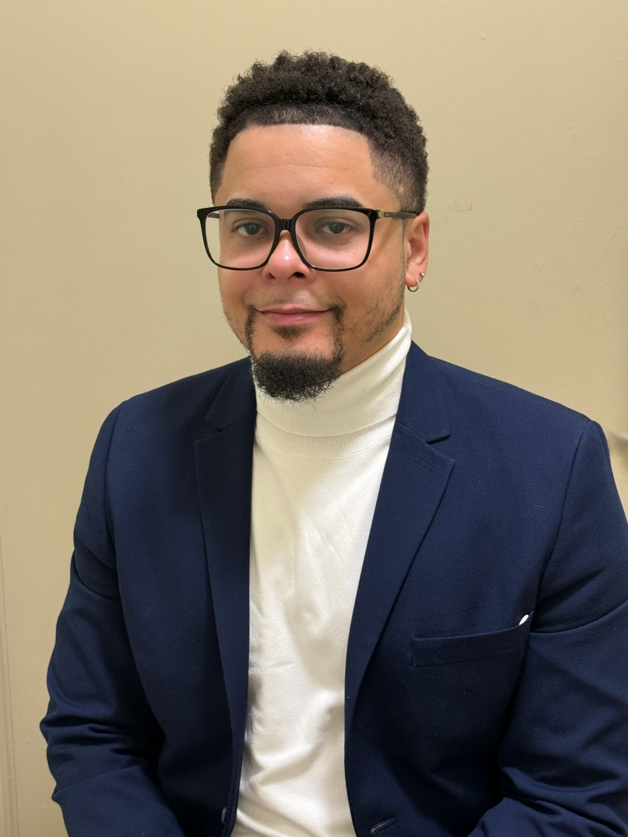A man with glasses, a goatee, and wearing a white turtleneck and navy blazer, sitting against a plain beige wall.