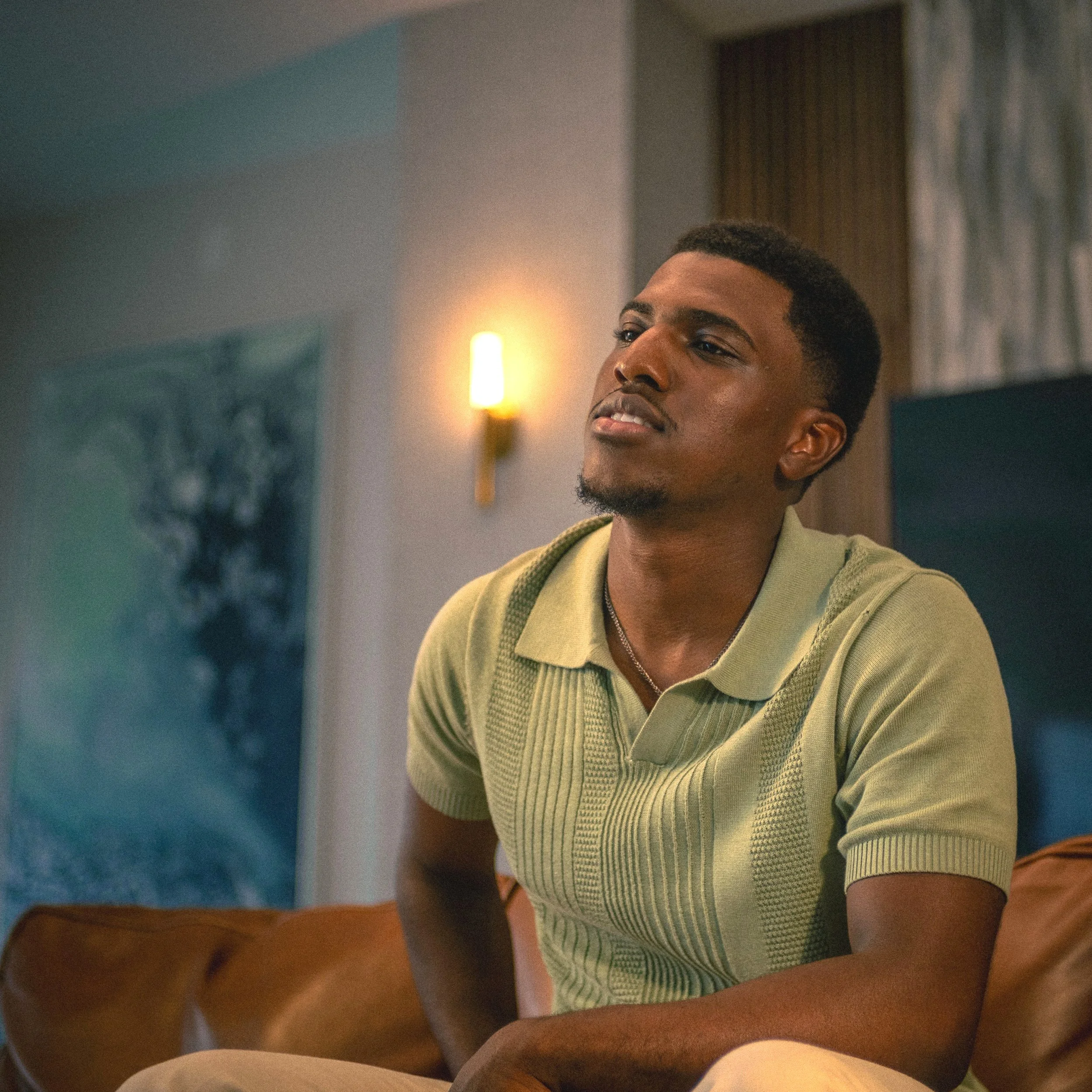 A man wearing a light green knit shirt sitting on a brown leather couch in a well-lit room with modern decor for portrait photography.