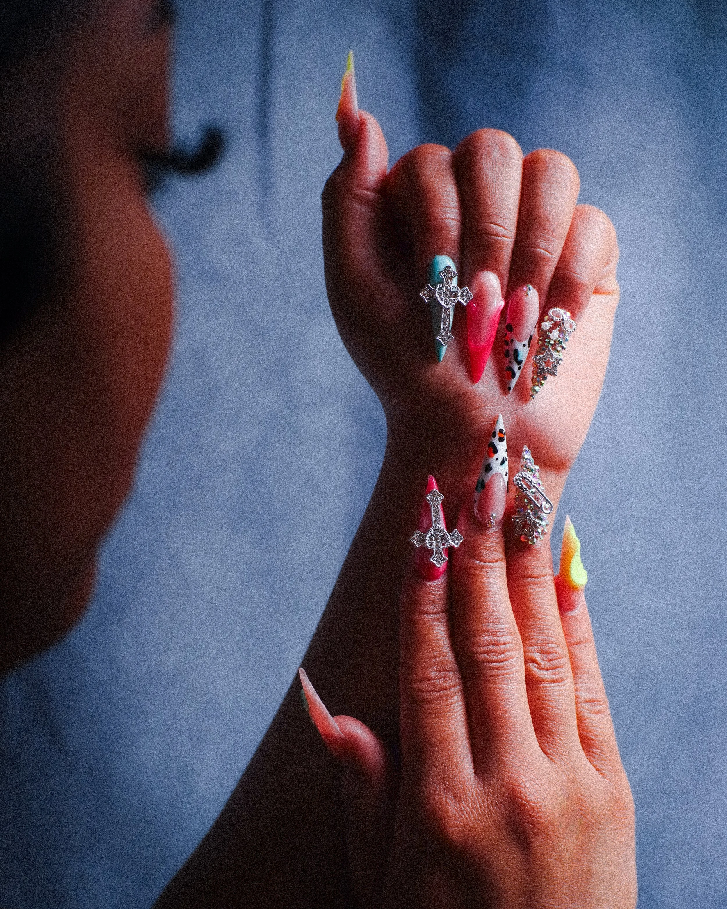 Close-up of a person's hands showcasing creative nail art with colorful rhinestones and designs on long, manicured nails against a blurred background for nail photography.