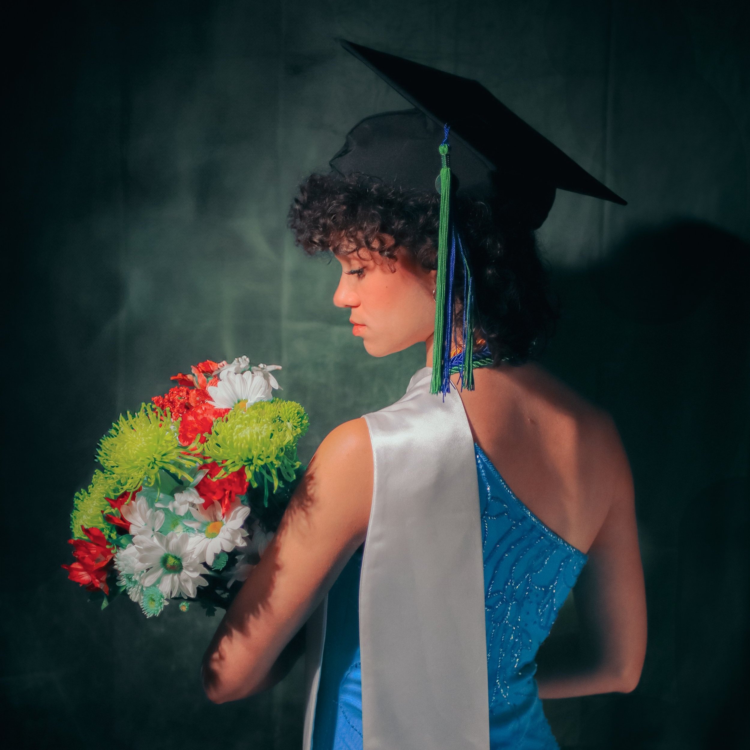Graduate in cap and gown holding a bouquet of flowers, wearing a blue dress with a white sash for a graduation photoshoot.