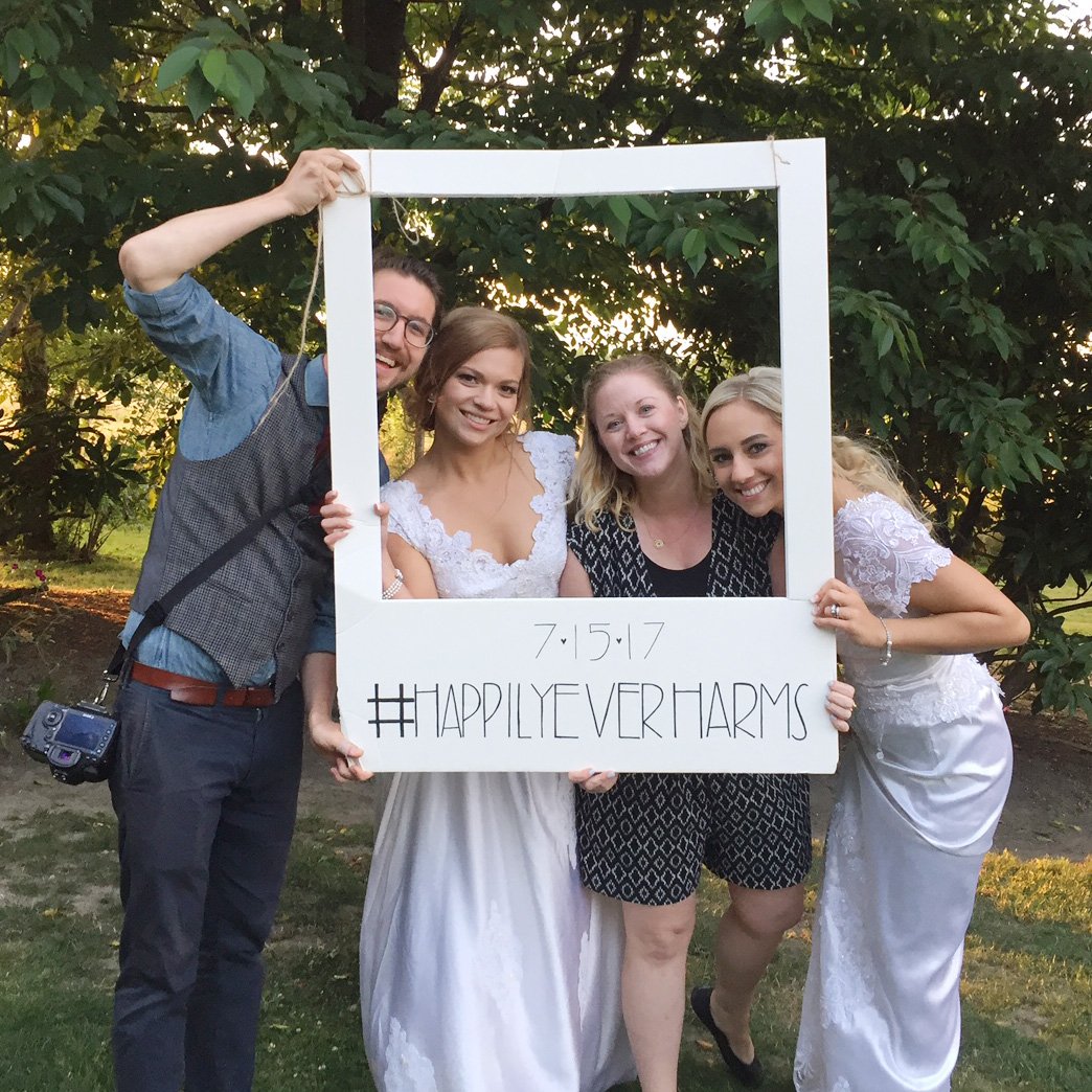 Group of four people posing outdoors with a photo frame prop displaying the date 7/15/17 and hashtag #HAPPILYEVERHARMS. Two individuals are wearing wedding dresses.