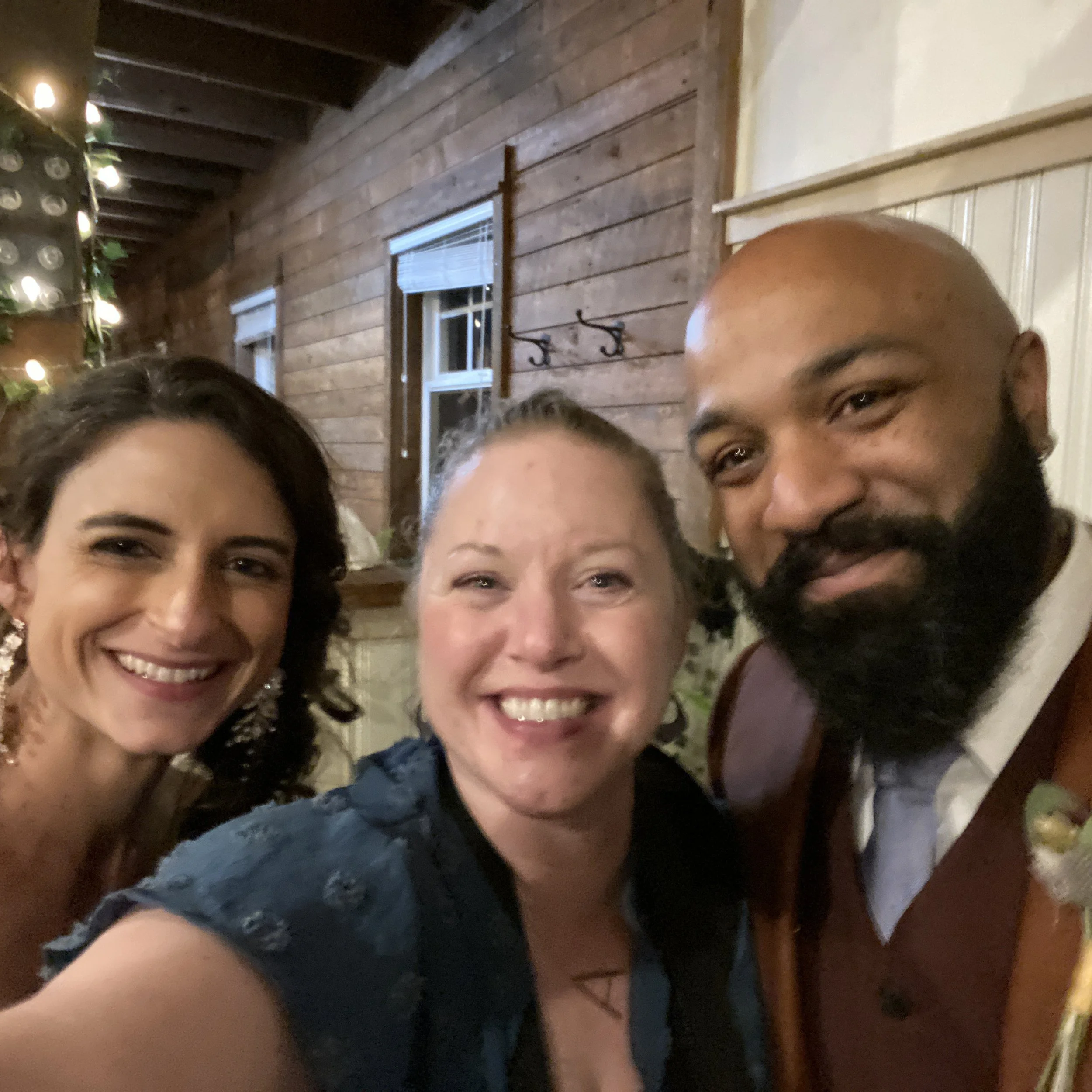 Three people smiling at a gathering, with wooden walls in the background.