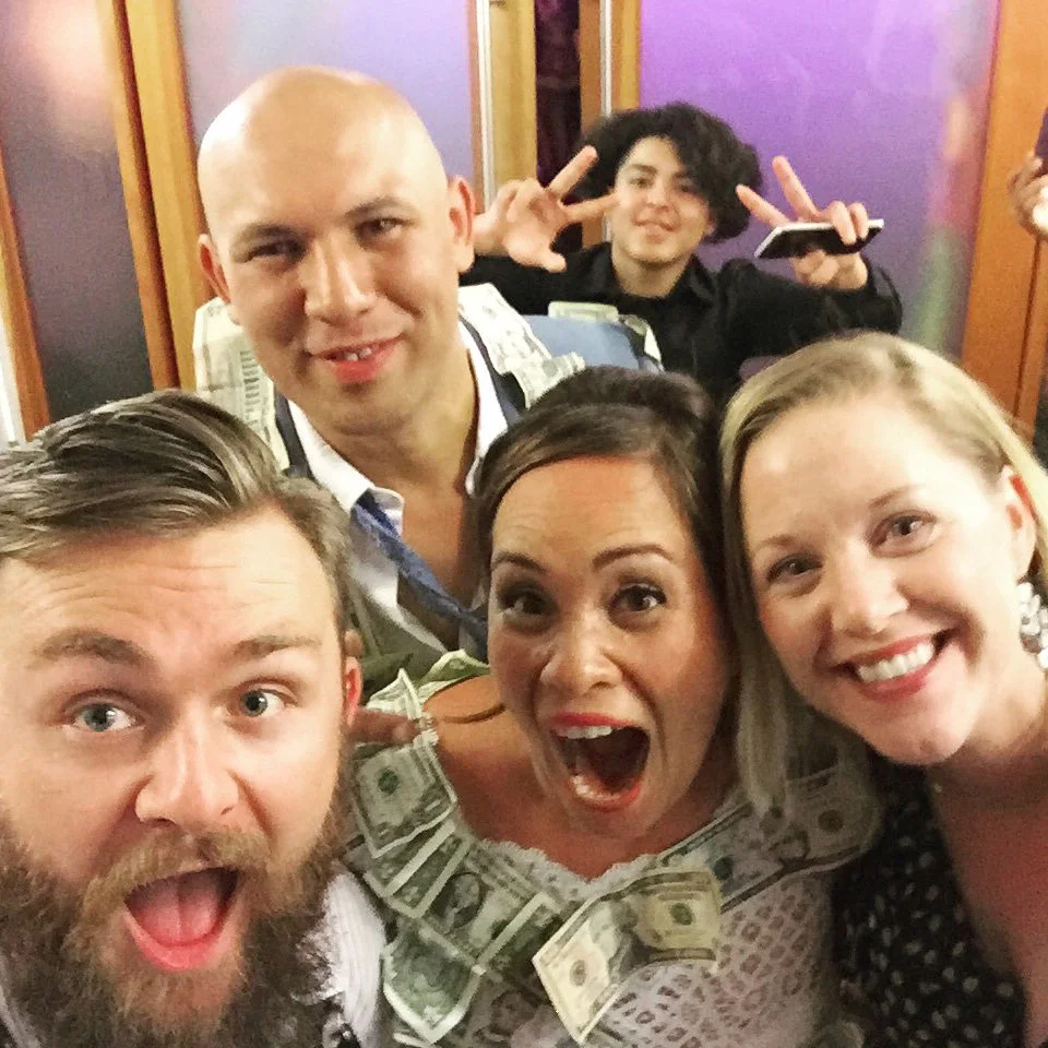 Group of five people posing happily, with one person wearing money pinned to their clothing, in a celebratory setting with a purple background. Another person is making a peace sign in the back.