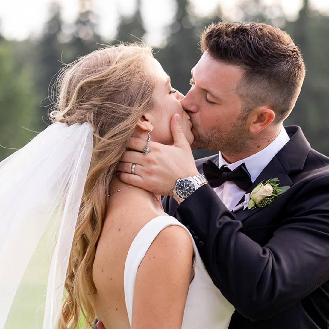 A bride and groom share a kiss outdoors during their wedding, with a blurred natural background.
