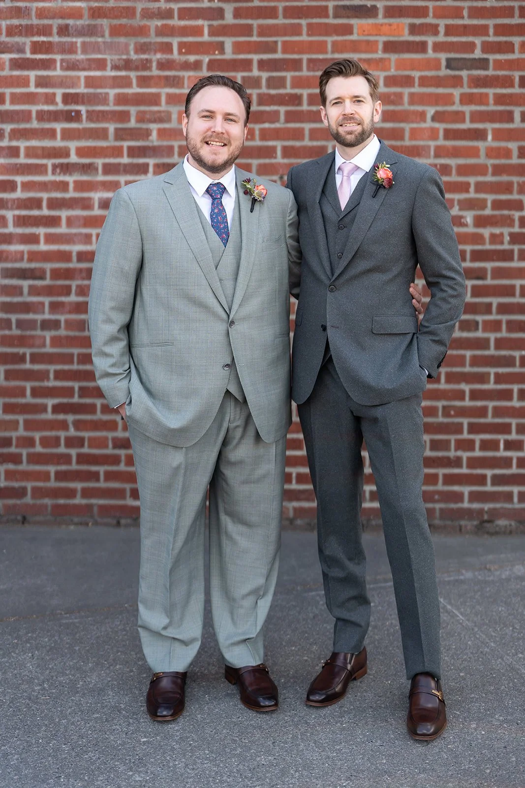 Two men in gray suits standing arm in arm against a brick wall, smiling at the camera.