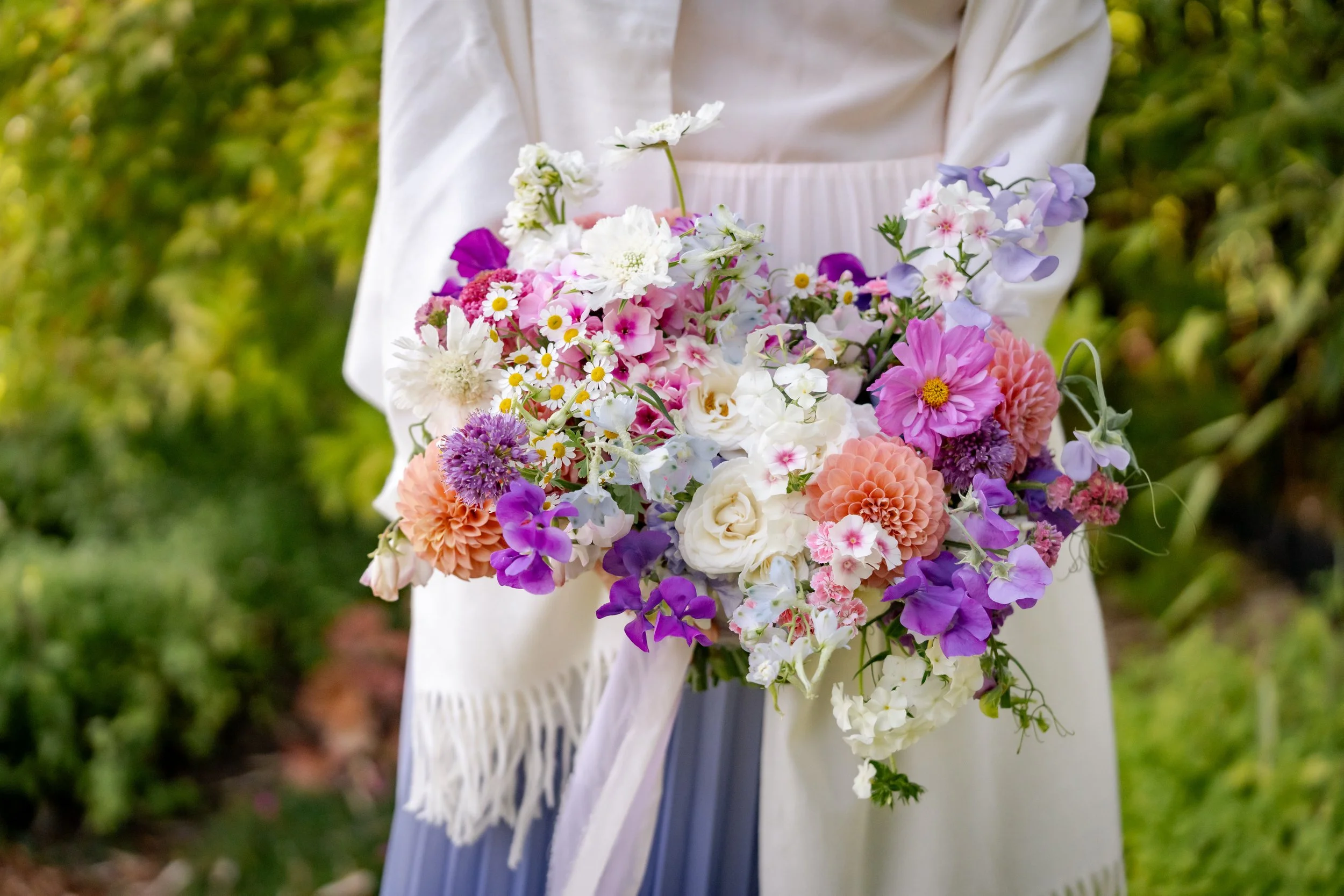 Person holding a colorful bouquet of various flowers, including roses, daisies, and dahlias, in an outdoor garden setting.