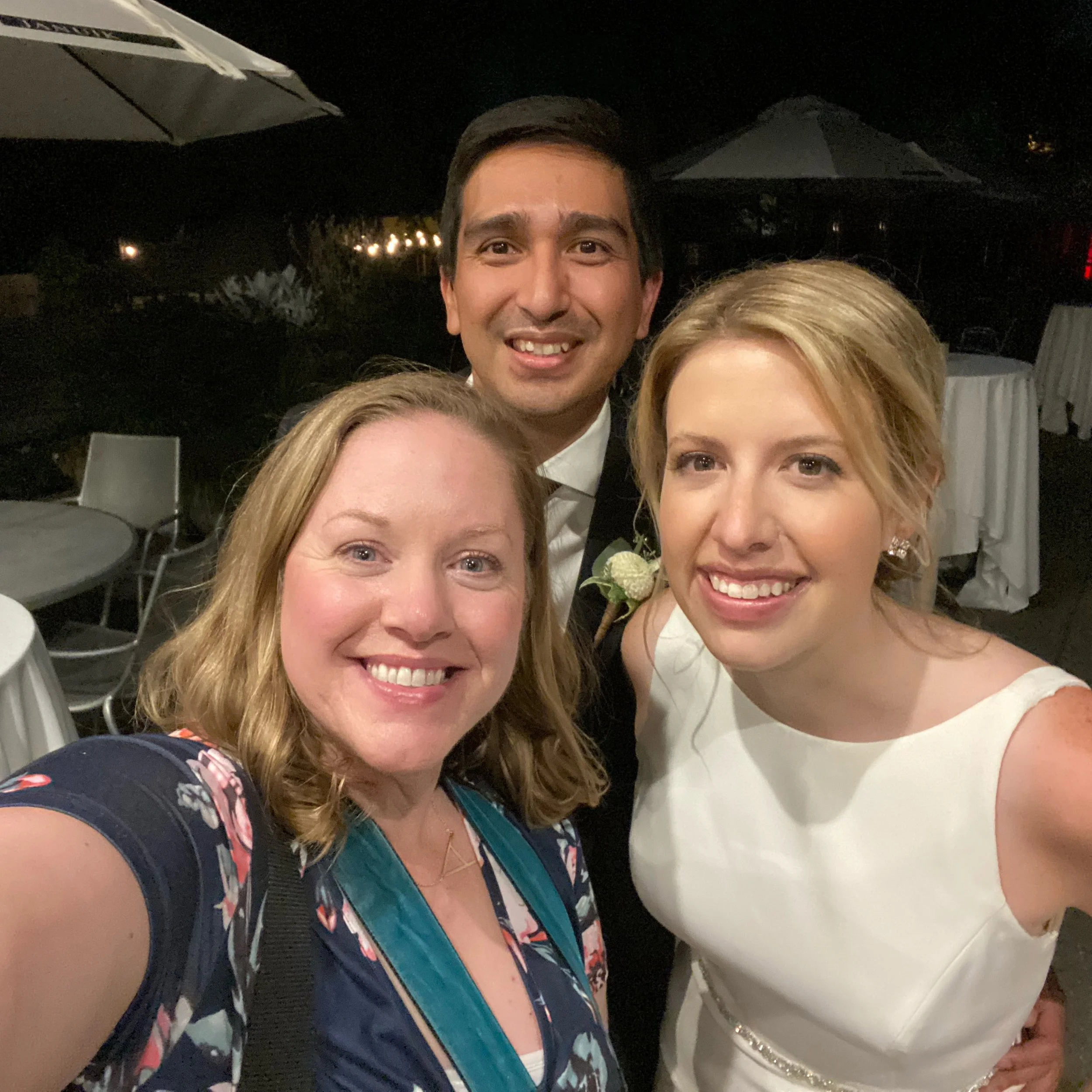 A smiling group selfie of three people, including a bride in a white dress, taken at an outdoor event with tables and umbrellas in the background.