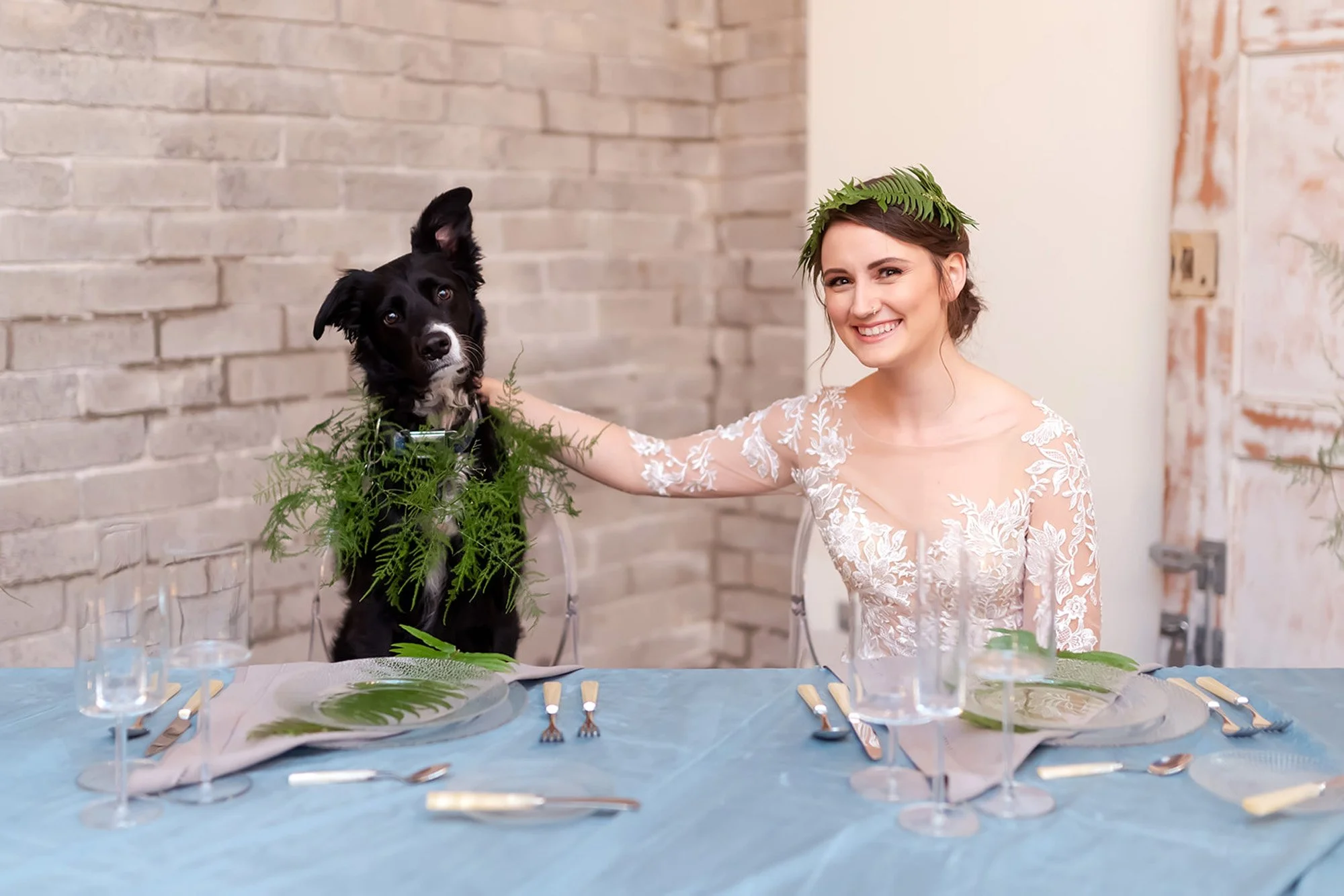 A bride in a lace wedding dress sits beside her black and white dog, both adorned with greenery, at a beautifully set table during her wedding day at the dog-friendly Within Sodo venue.
