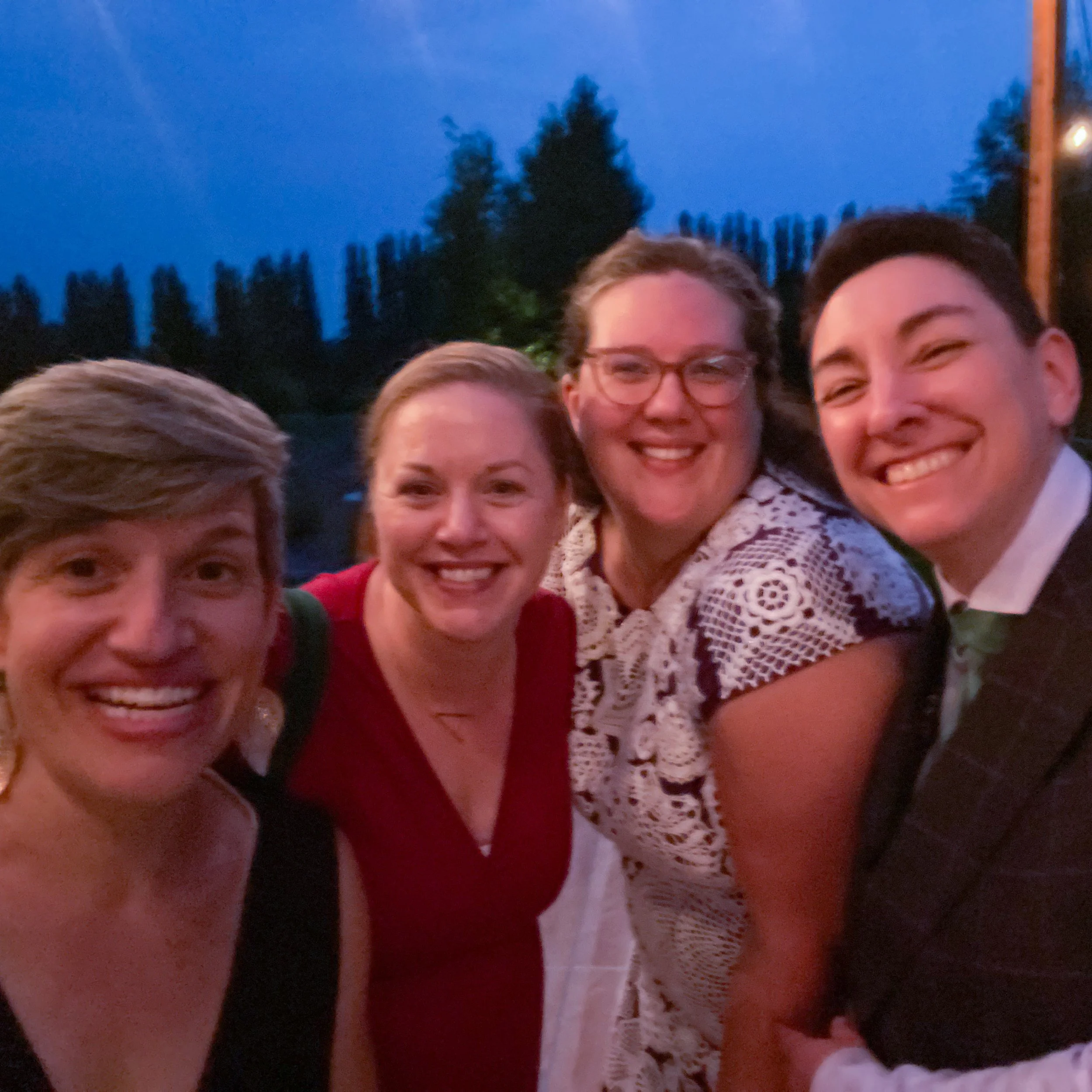 A group of four adults smiling outdoors at dusk, with trees in the background. They appear to be dressed for a formal event.