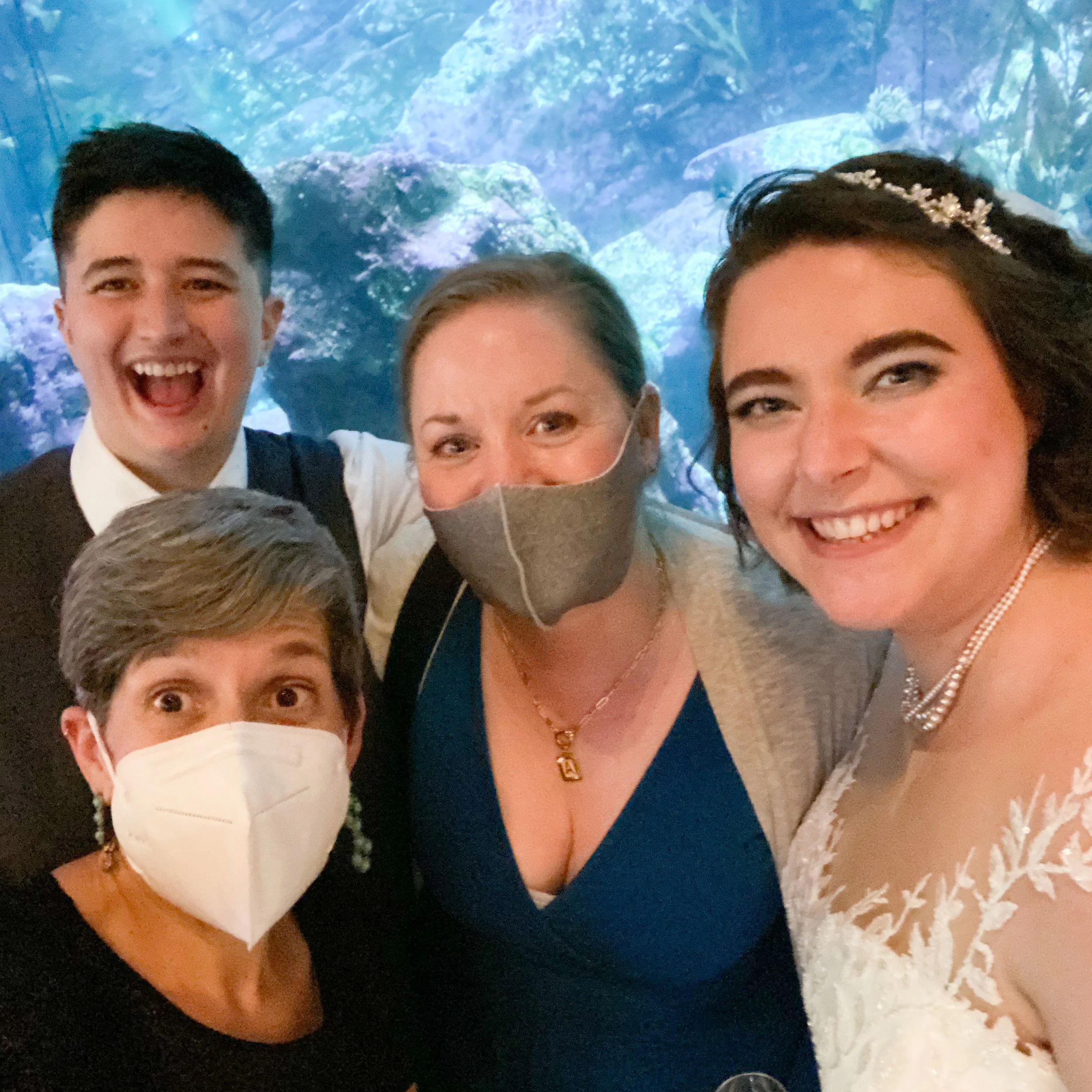 Group of people smiling and wearing masks, one in wedding attire, in front of aquarium backdrop.