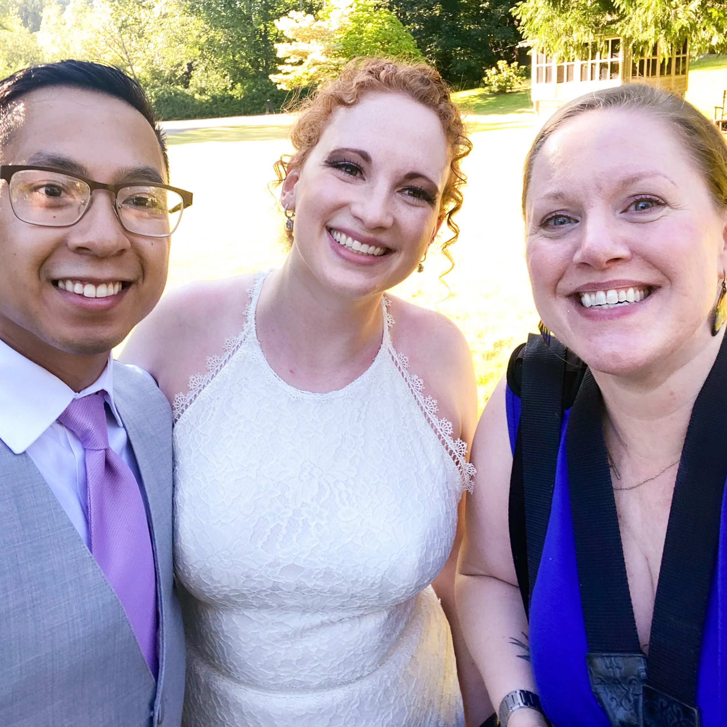 Three people smiling outdoors in a sunny setting. One person is in a wedding dress, another in a suit with a pink tie, and the third in a blue dress with a camera strap.
