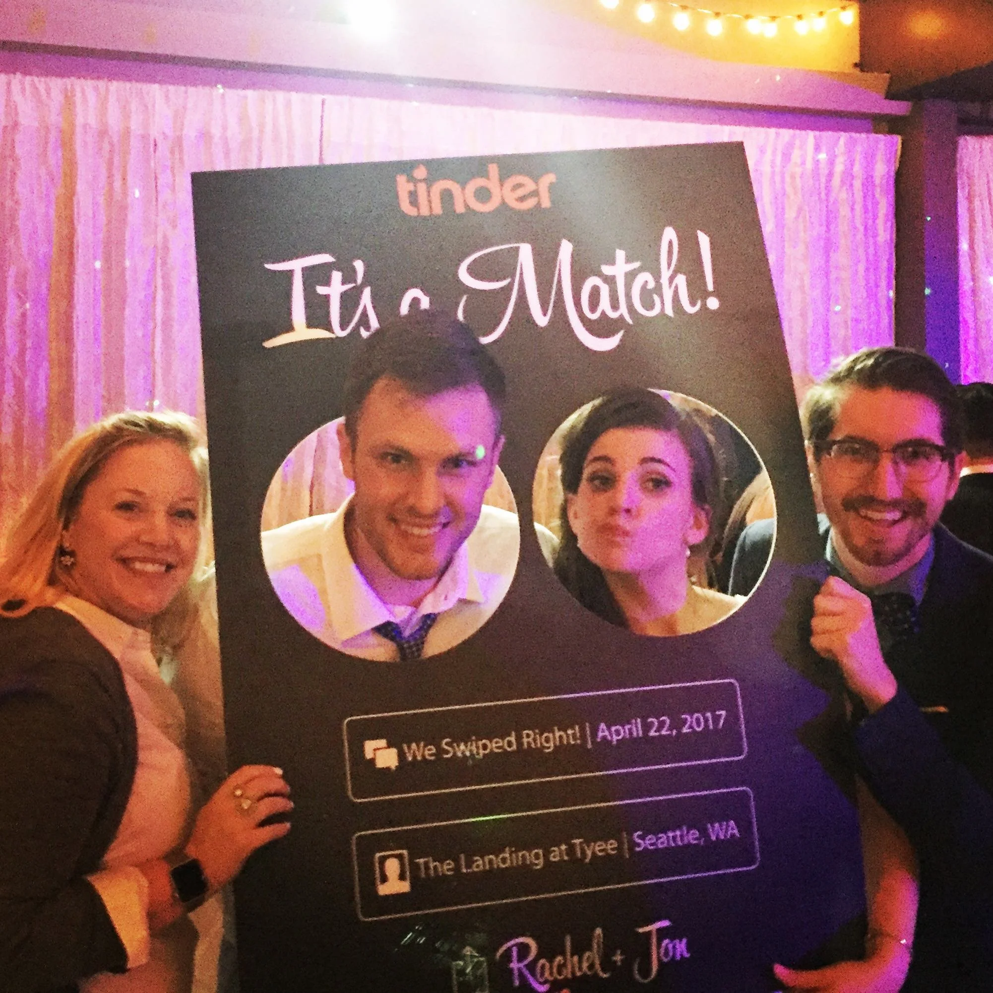 Group of people posing with large "It's a Match!" Tinder-themed sign at an event, displaying date and location details.