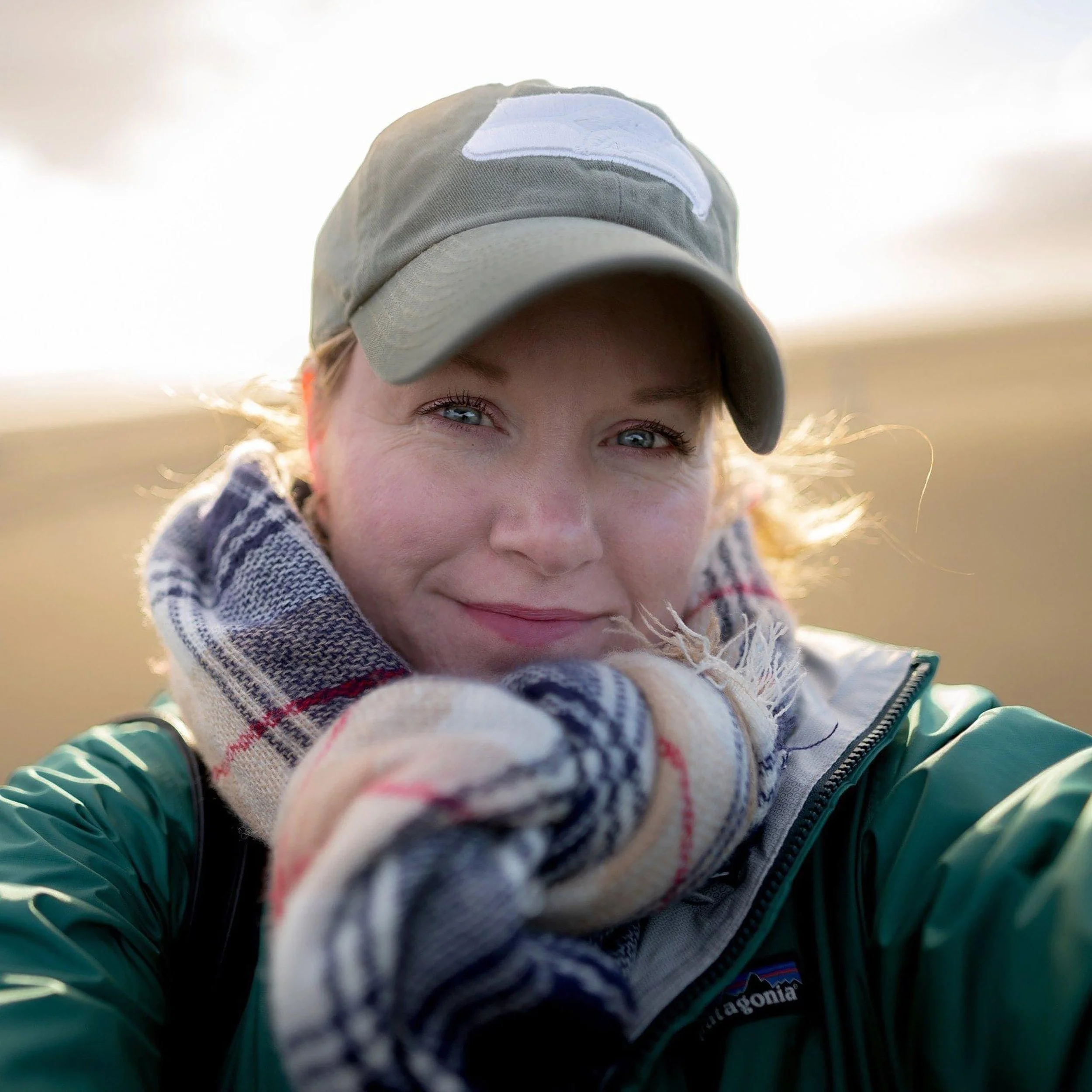 Close-up of a woman outdoors wearing a green jacket, a plaid scarf, a gray baseball cap, and gloves, smiling as the sunlight shines on her face.