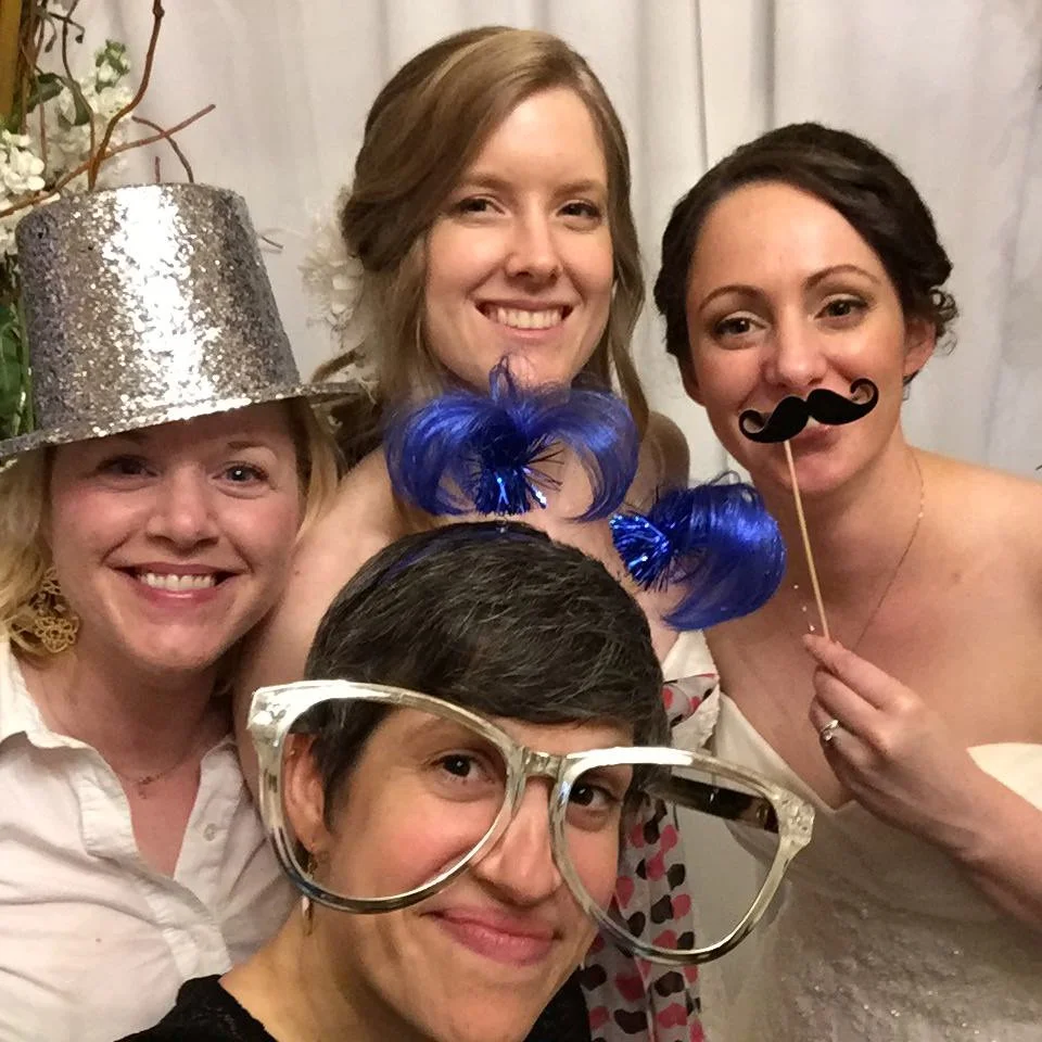 Group of four people smiling in a photo booth, wearing novelty accessories like a silver glitter hat, large glasses, blue feather headband, and fake mustache on a stick.