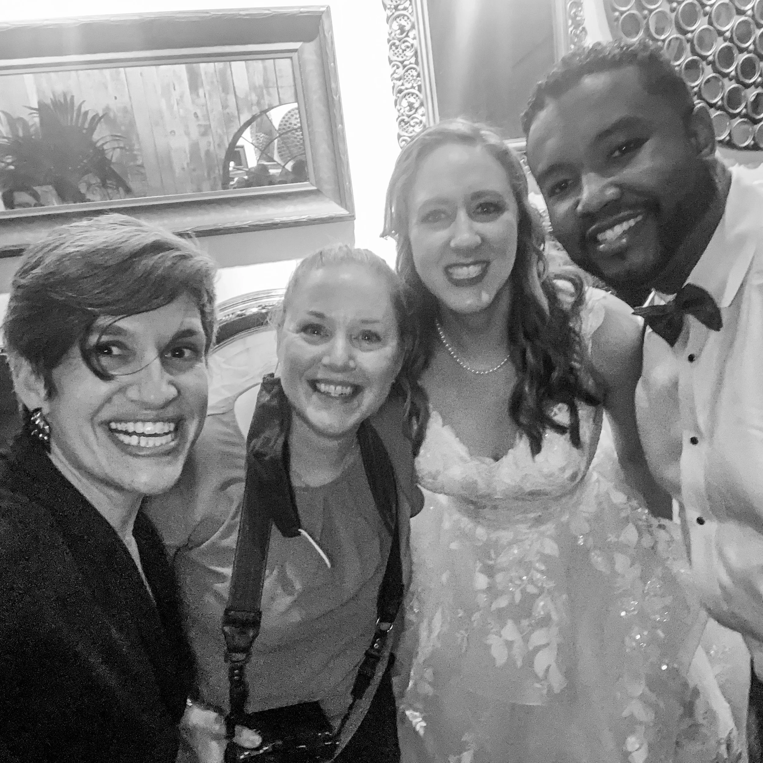 A black and white photo of four smiling adults, including a bride in a wedding dress and a groom in a tuxedo, with two other guests posing closely. The background features decorative wall mirrors.
