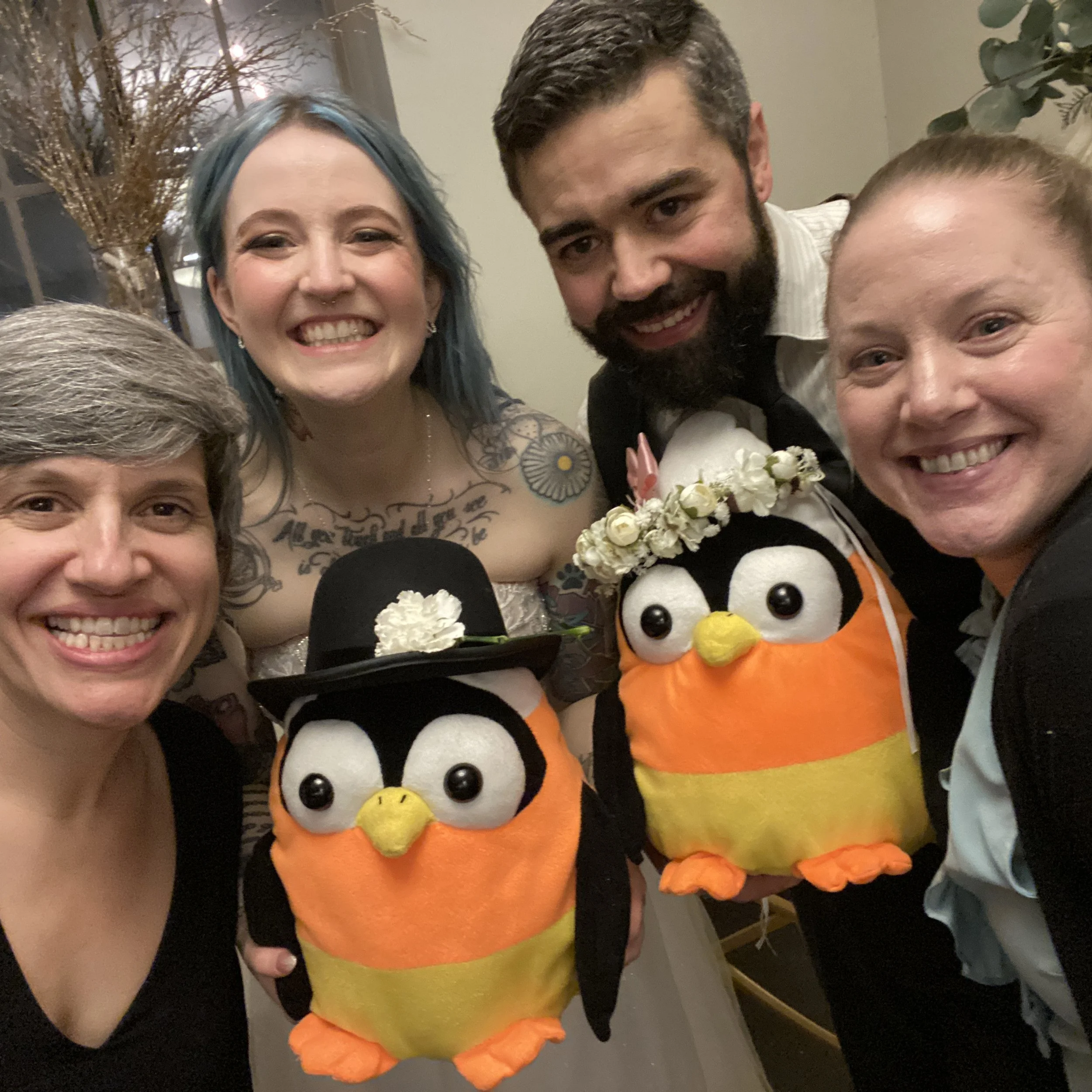 Group smiling with two stuffed penguins dressed in wedding attire, one with a black top hat and the other with a flower crown, inside a room with a plant.