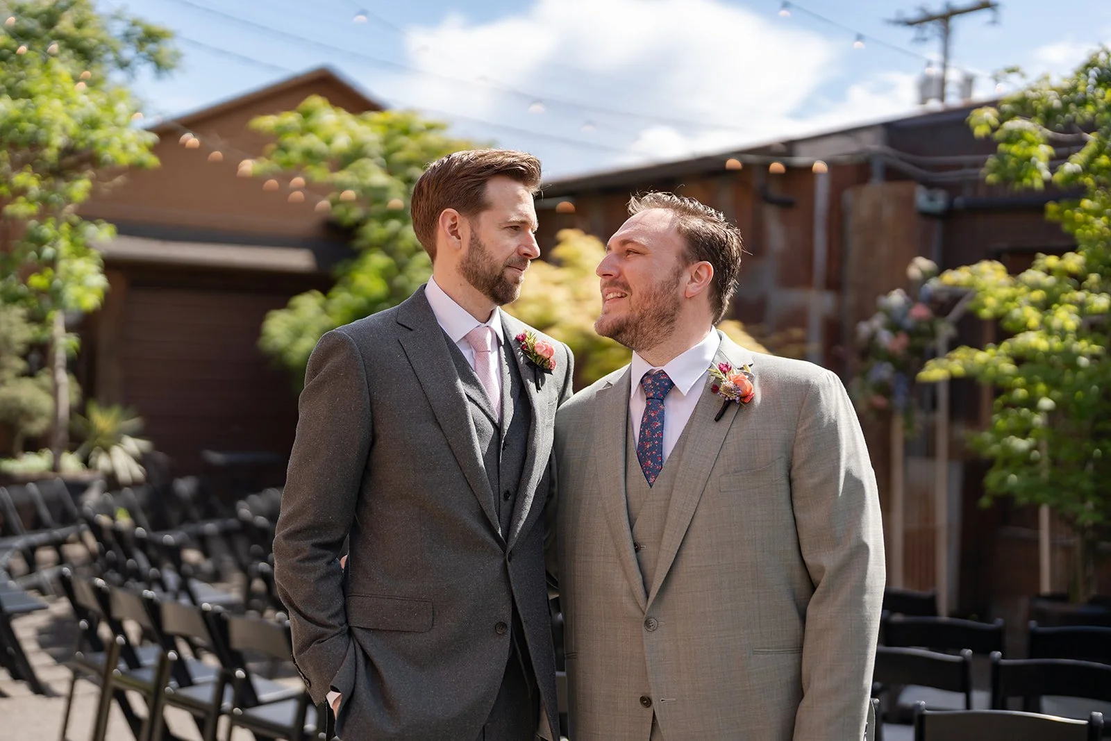 Two men dressed in suits with boutonnières, standing close and looking at each other at an outdoor wedding ceremony.