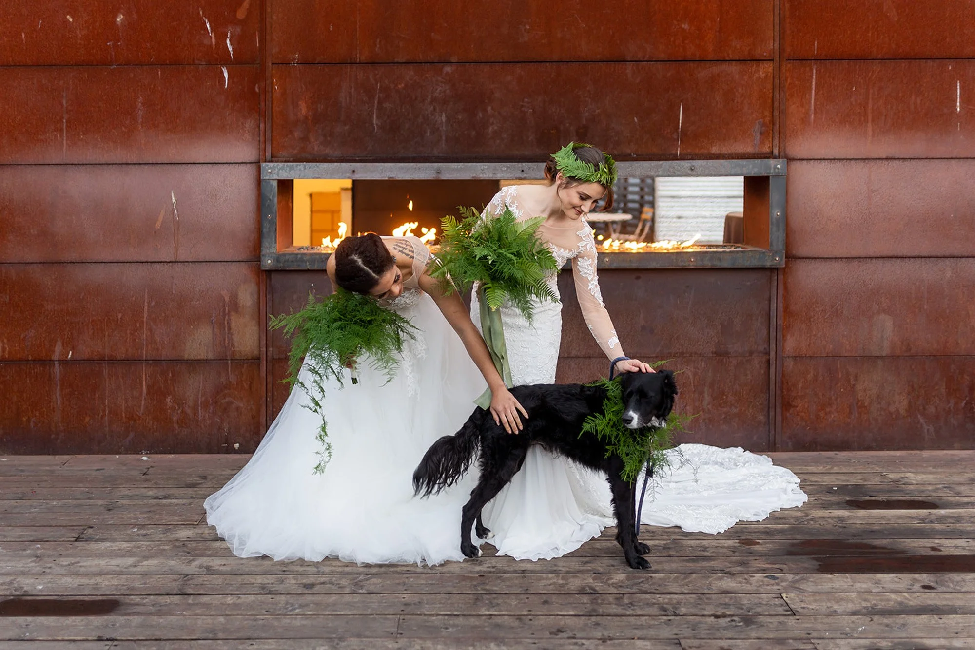 Two brides in white wedding dresses, adorned with greenery, lovingly pet a black dog decorated with matching foliage on the rooftop deck of Within Sodo in Seattle, with a rustic metal wall and open fire feature in the background.