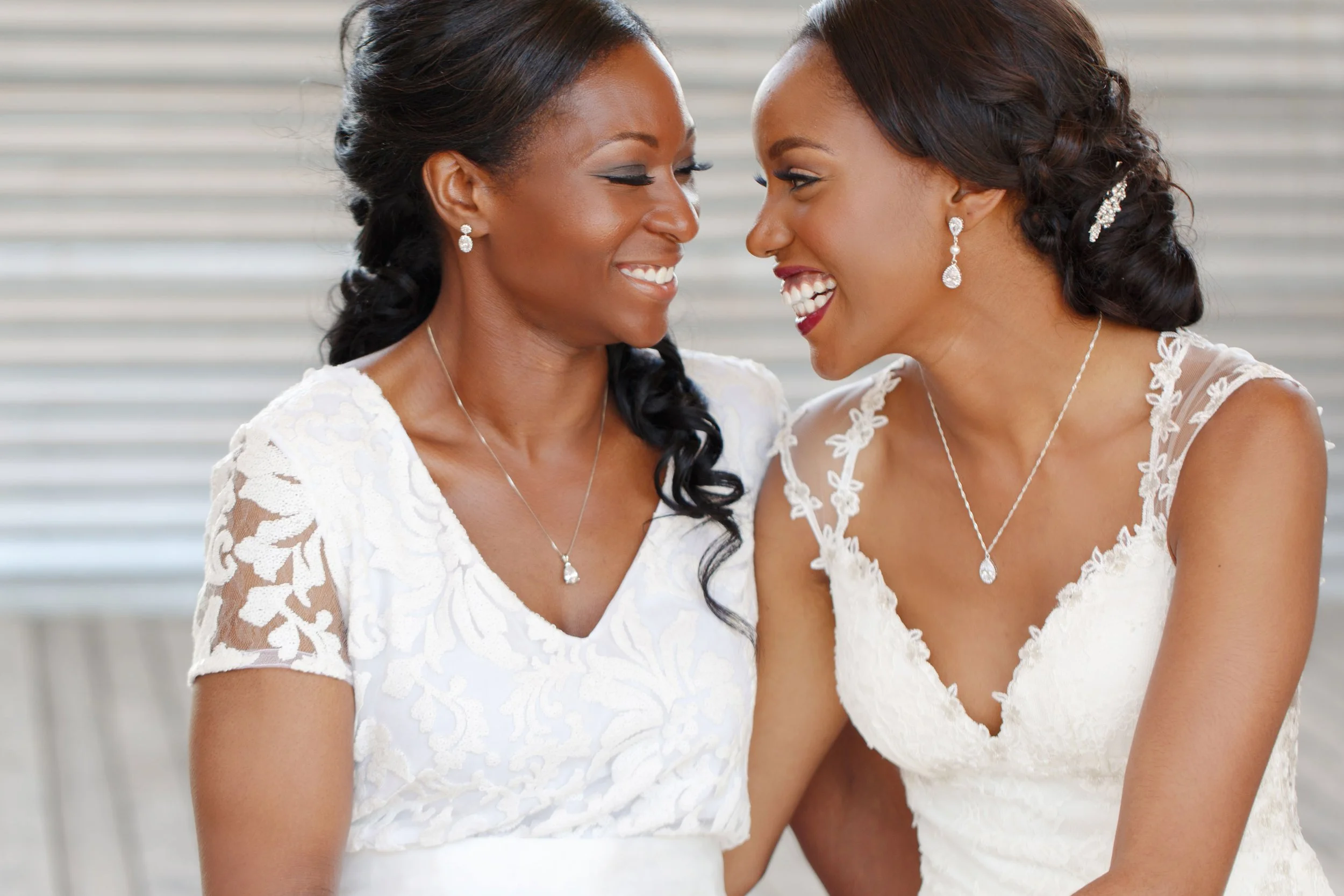 Two women dressed in white wedding gowns sharing a joyful moment, smiling and touching foreheads.