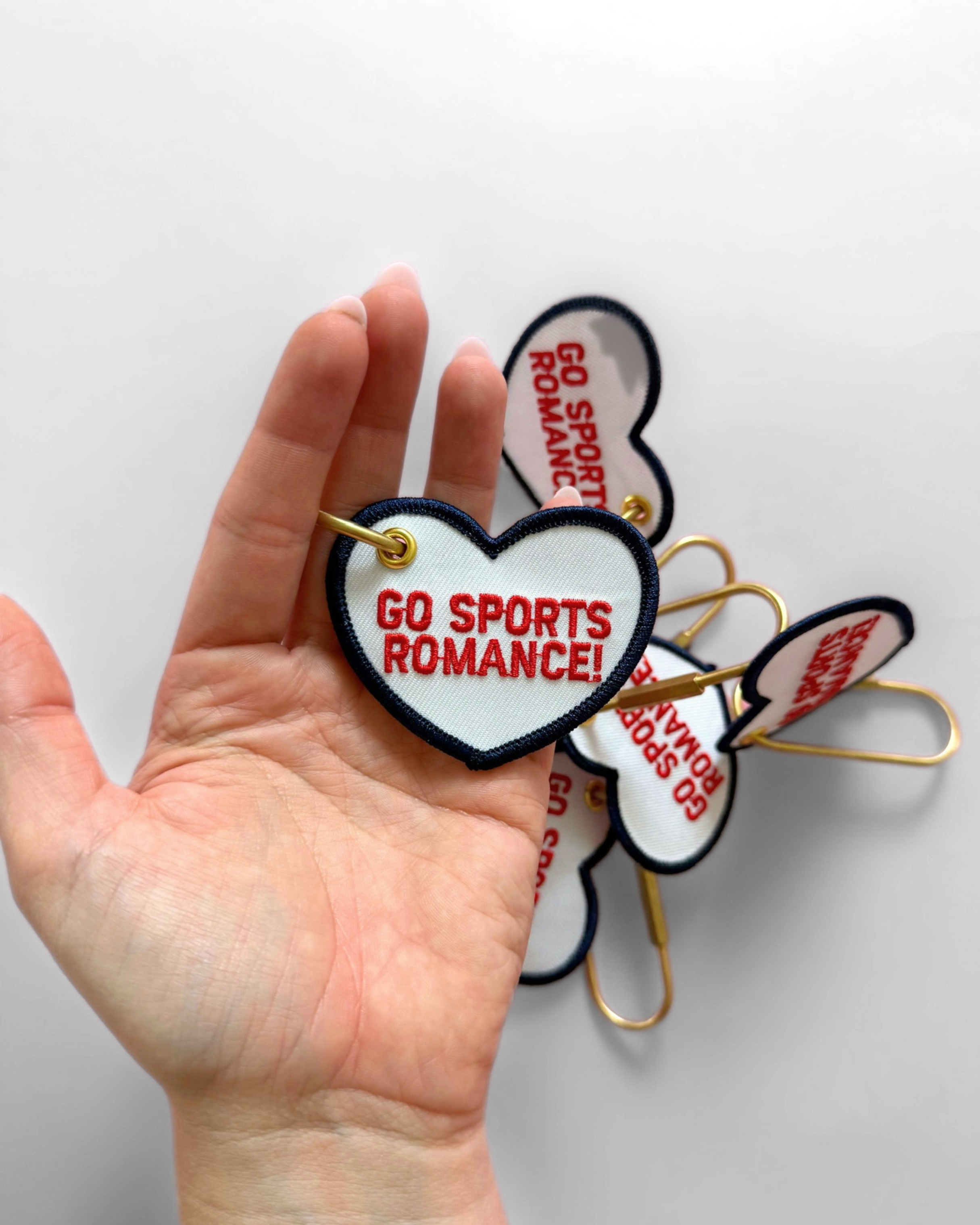 White heart-shaped embroidered keychains with navy trim and red “Go Sports Romance!” lettering, finished with oversized gold safety-pin style hardware.