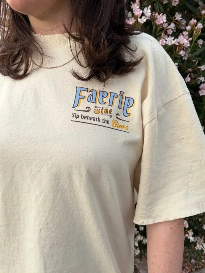 Back view of a model wearing an oversized ivory T-shirt with a blue, gold, and dark brown stained-glass-style graphic reading “Faerie Wine” and “Sip beneath the Stars."