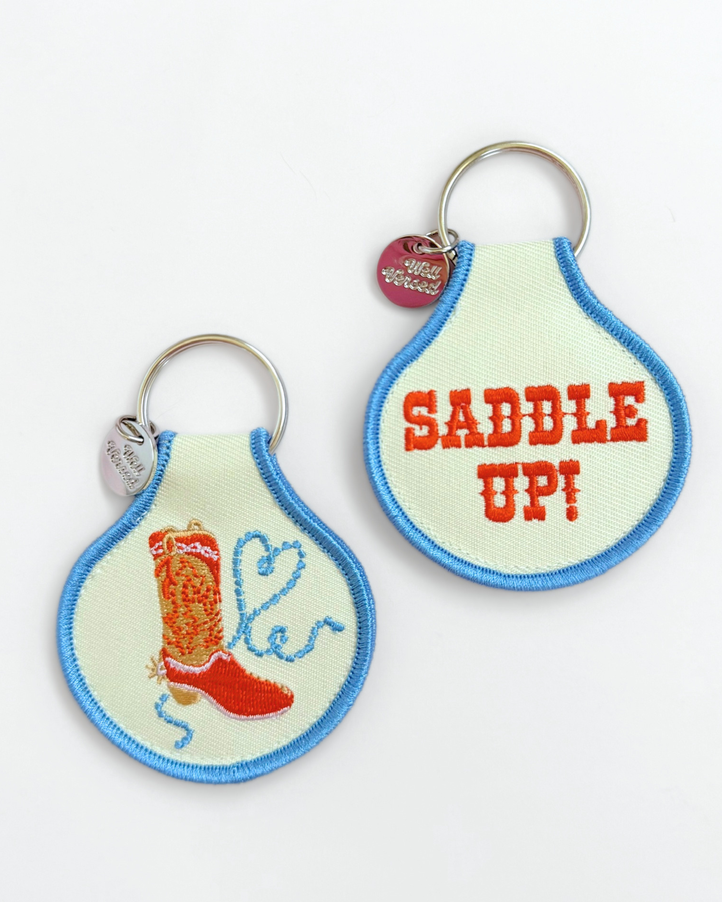 Double-sided embroidered fabric keychain with blue trim, featuring a red cowboy boot and lasso on one side and “Saddle Up!” in orange Western lettering on the other.
