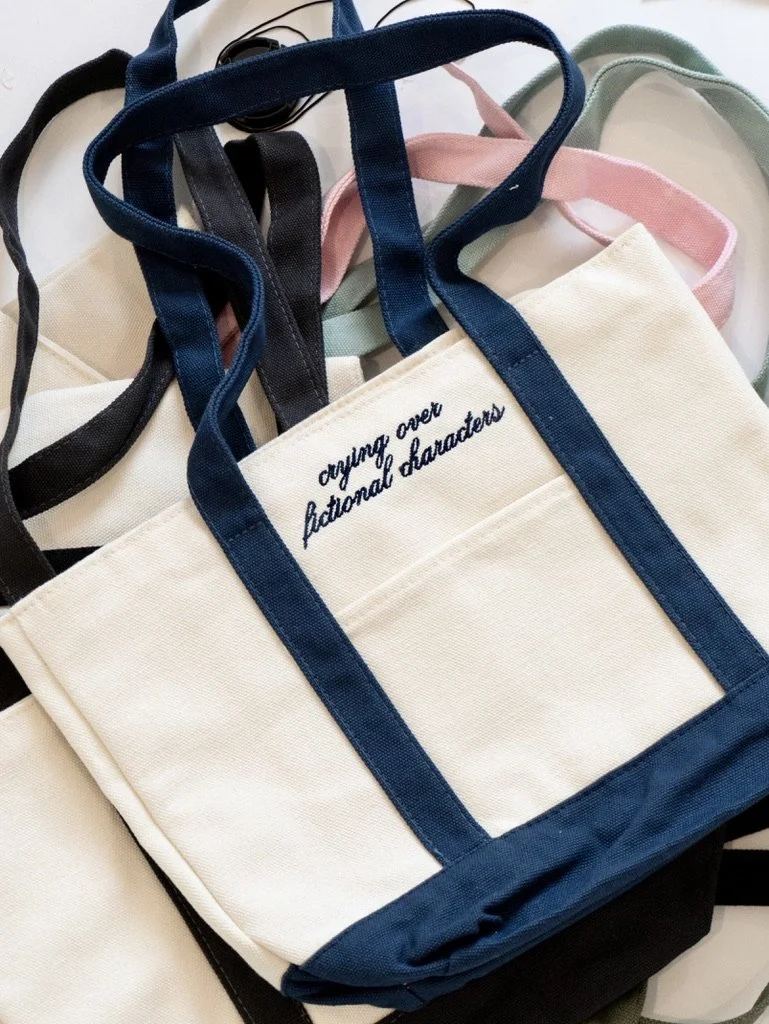 Crying Over Fictional Characters embroidered canvas tote bag with navy straps, a cute bookish tote for readers, romance book lovers, bookstore trips, and reader gifts.