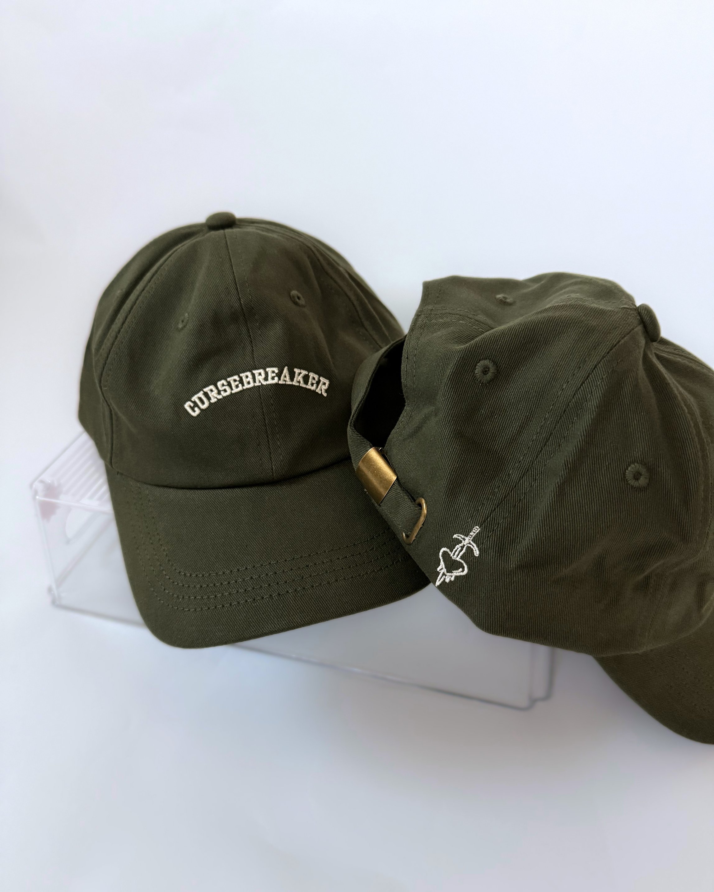 Two olive green baseball caps showing cream “CURSEBREAKER” embroidery on the front and a heart pierced by a sword beside an adjustable brass-tone clasp on the back.