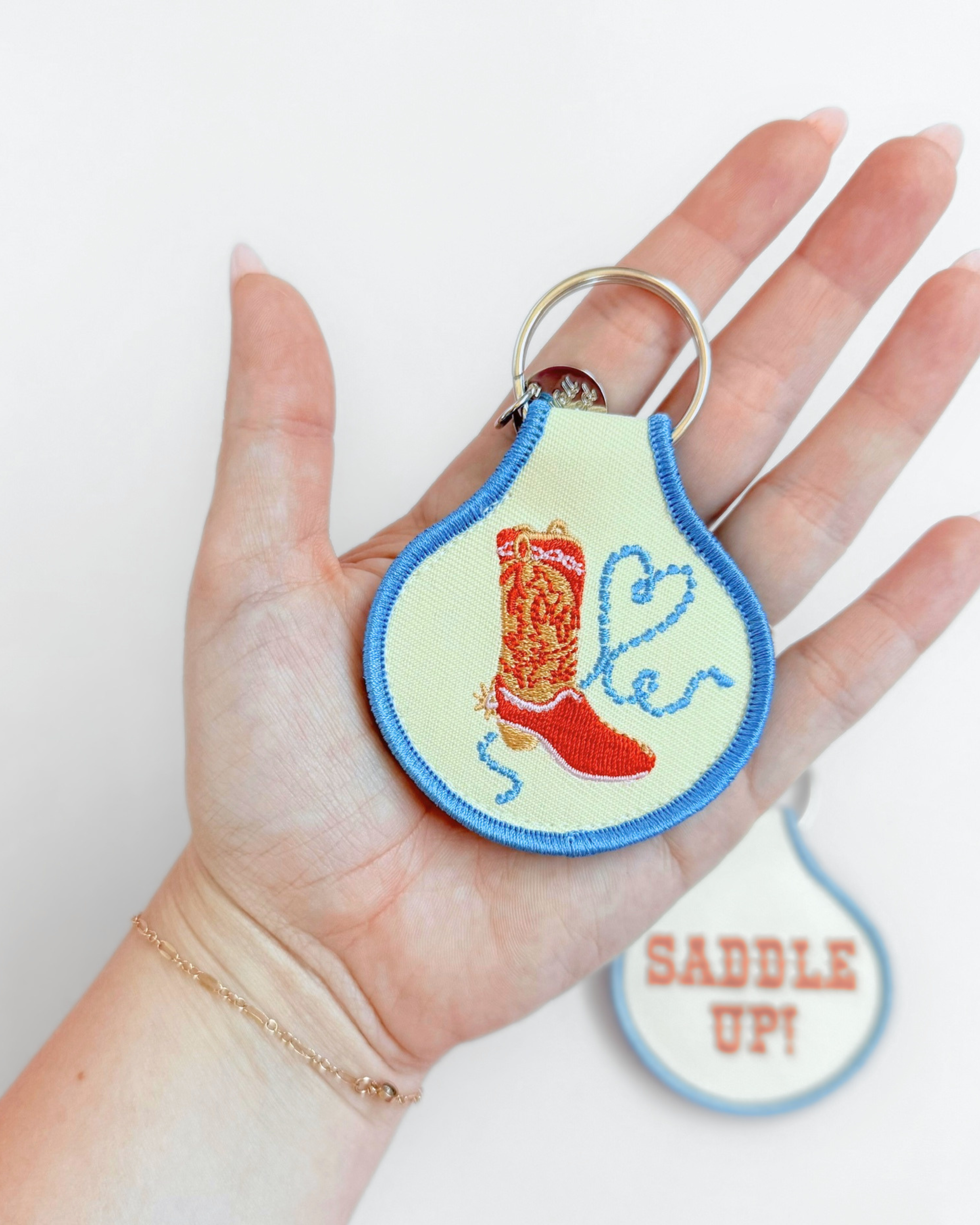 Double-sided embroidered fabric keychain with blue trim, featuring a red cowboy boot and lasso on one side and “Saddle Up!” in orange Western lettering on the other.