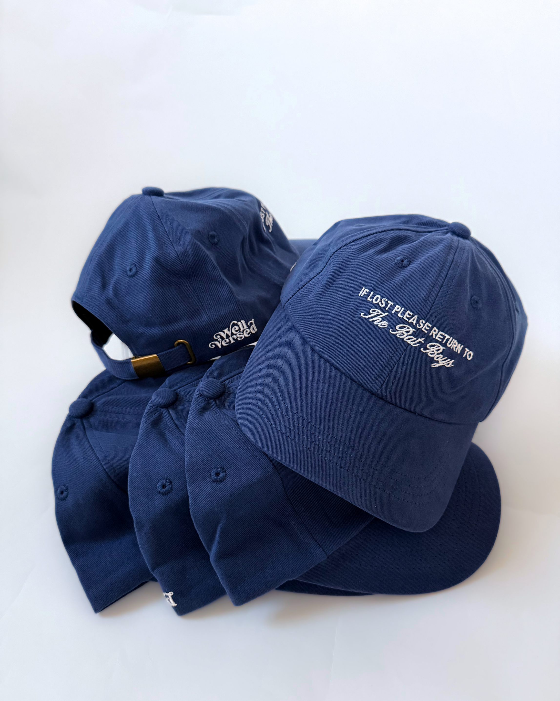 Stacked blue baseball caps with cream “If Lost Please Return to The Bat Boys” embroidery on the front, Well Versed branding on the back, and an adjustable brass-tone clasp.