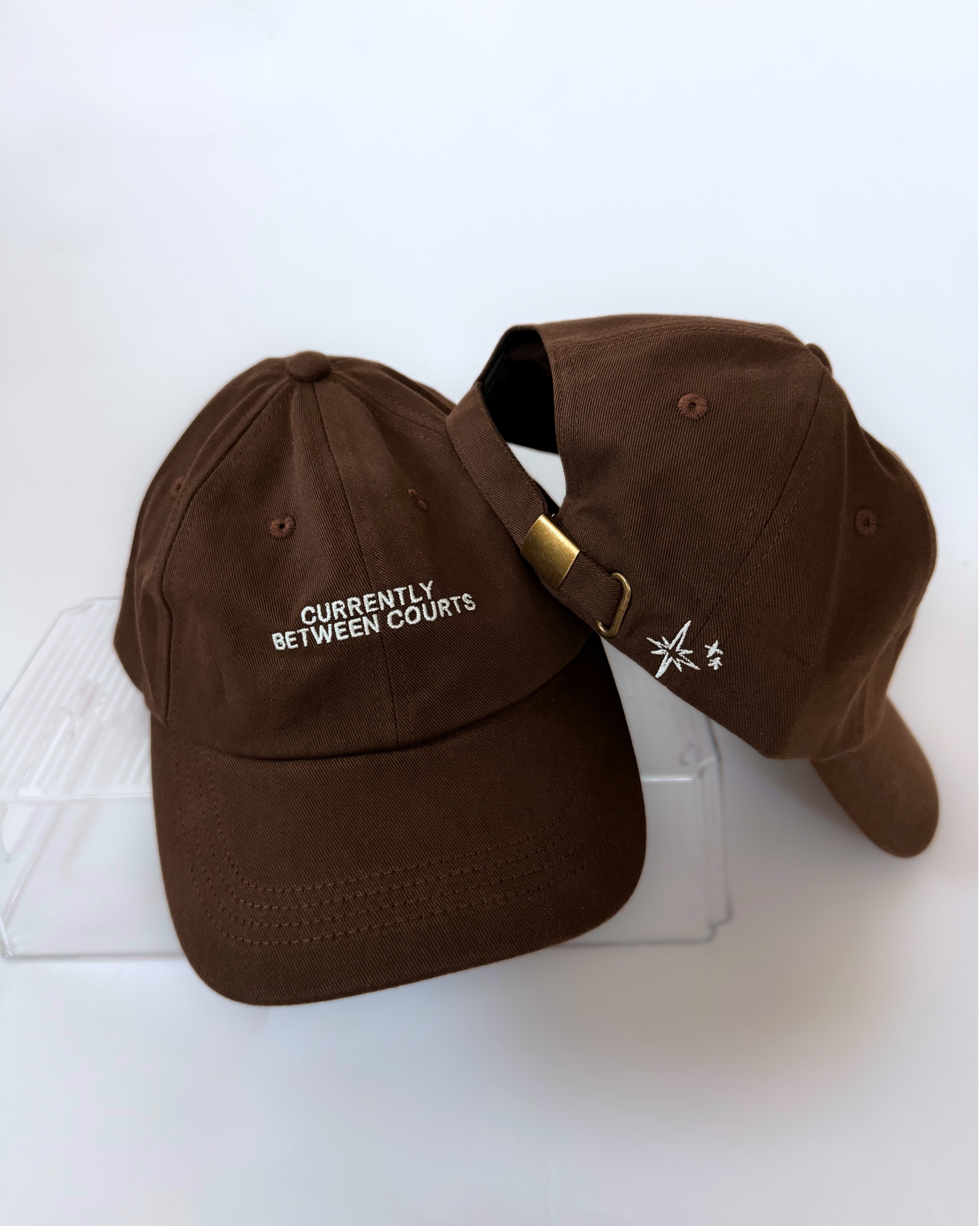 Two brown baseball caps showing cream “Currently Between Courts” embroidery on the front and embroidered stars beside an adjustable brass-tone clasp on the back.