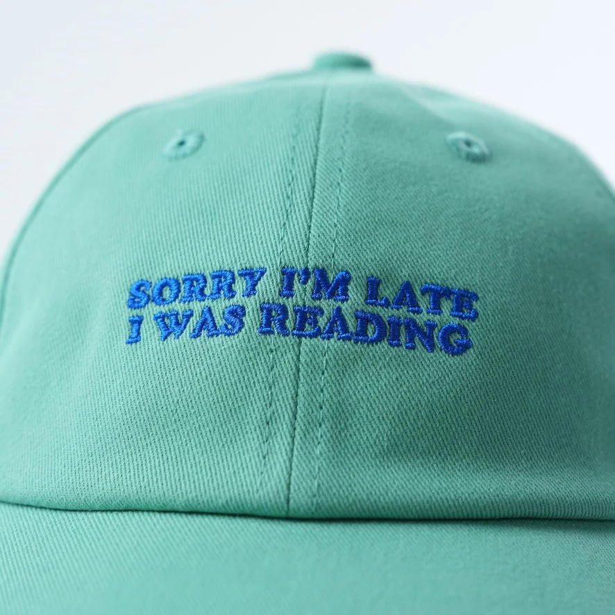 Mint green baseball cap embroidered with the phrase “SORRY I'M LATE I WAS READING” in bright blue lettering.
