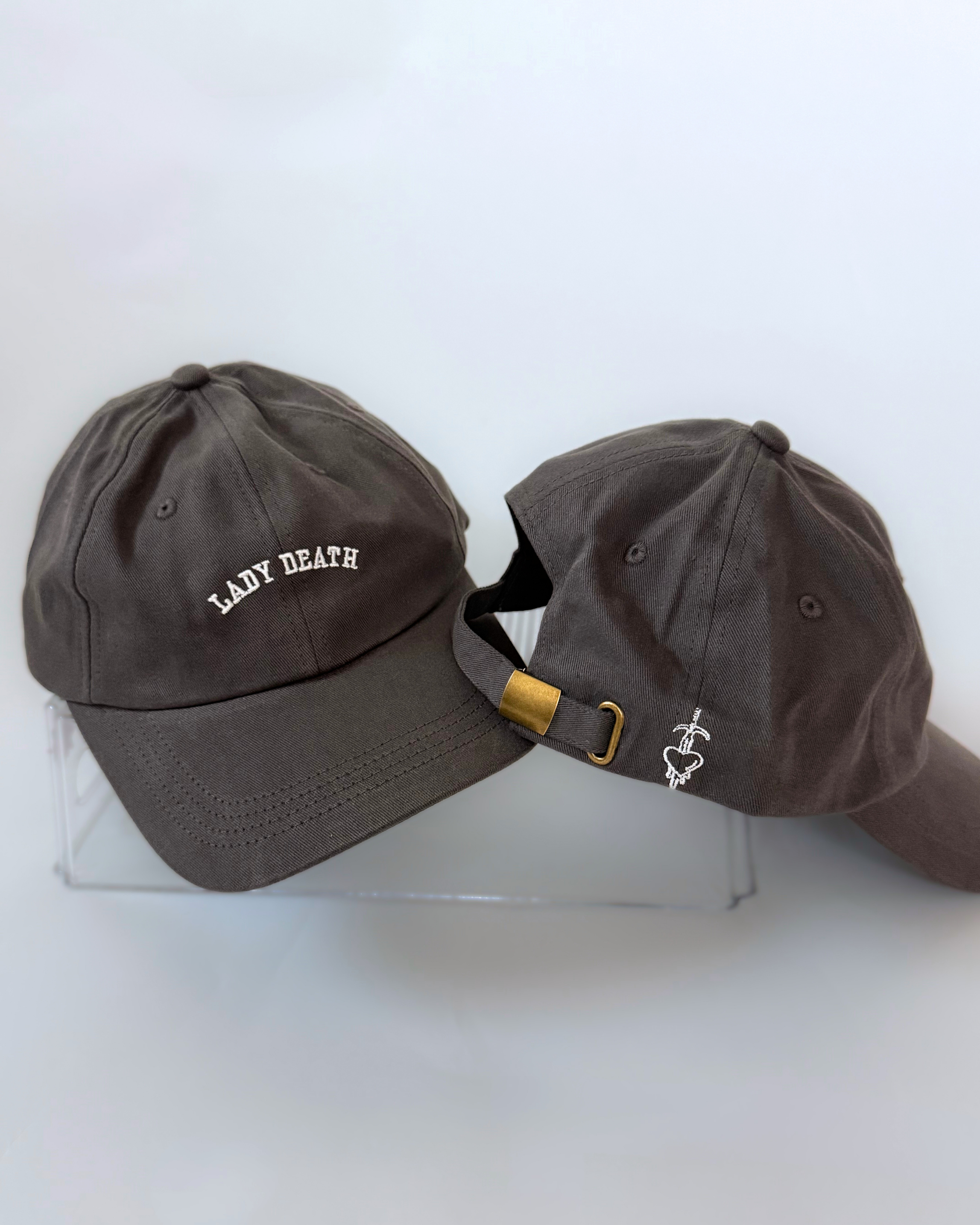 Two charcoal gray baseball caps showing cream “LADY DEATH” embroidery on the front and a heart pierced by a sword beside an adjustable brass-tone clasp on the back.