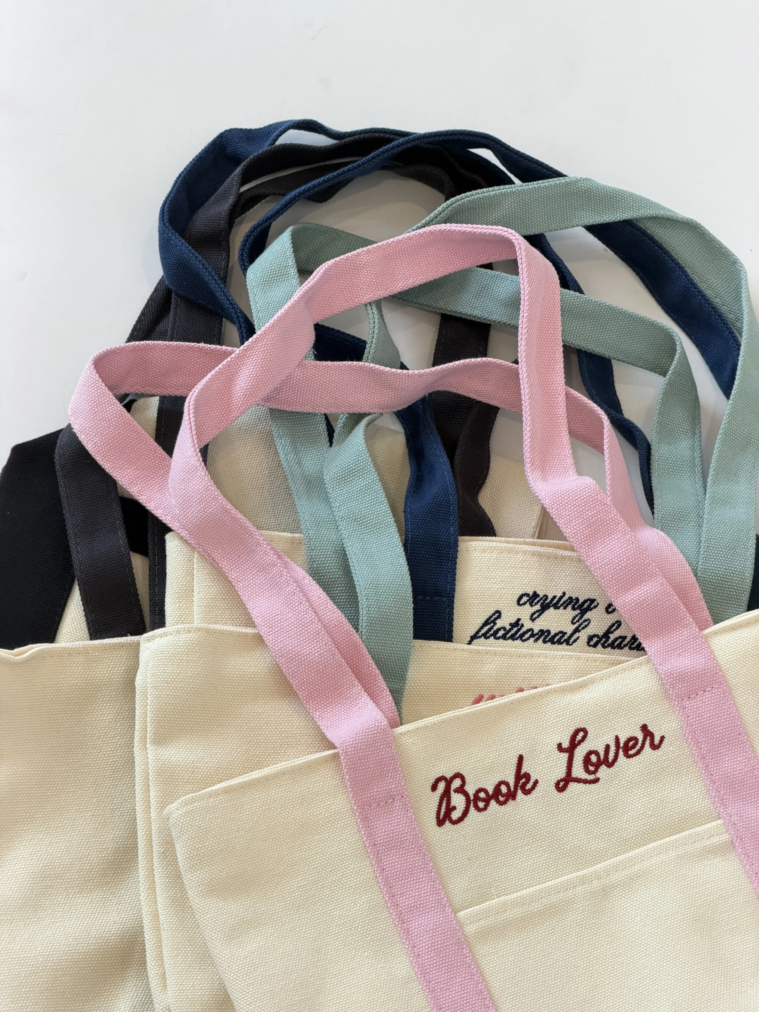 Stack of Tote backs featuring bookish phrases such as book lover and crying over fictional characters
