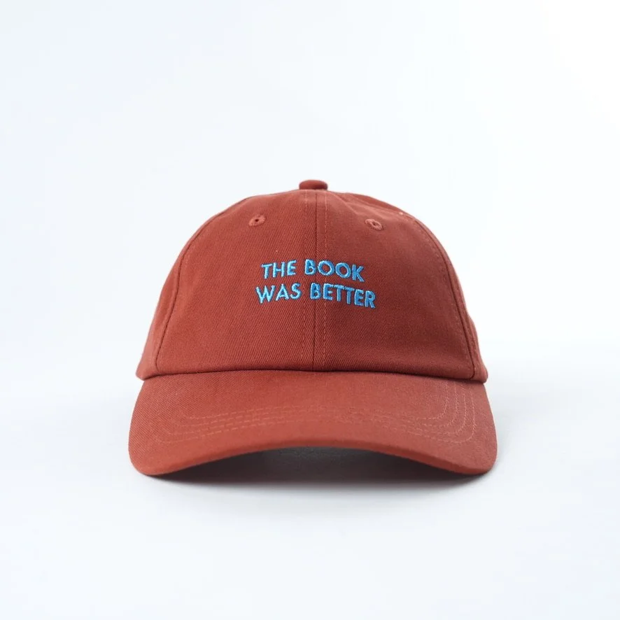 Rust-colored baseball cap embroidered with the phrase “THE BOOK WAS BETTER” in bright blue lettering.