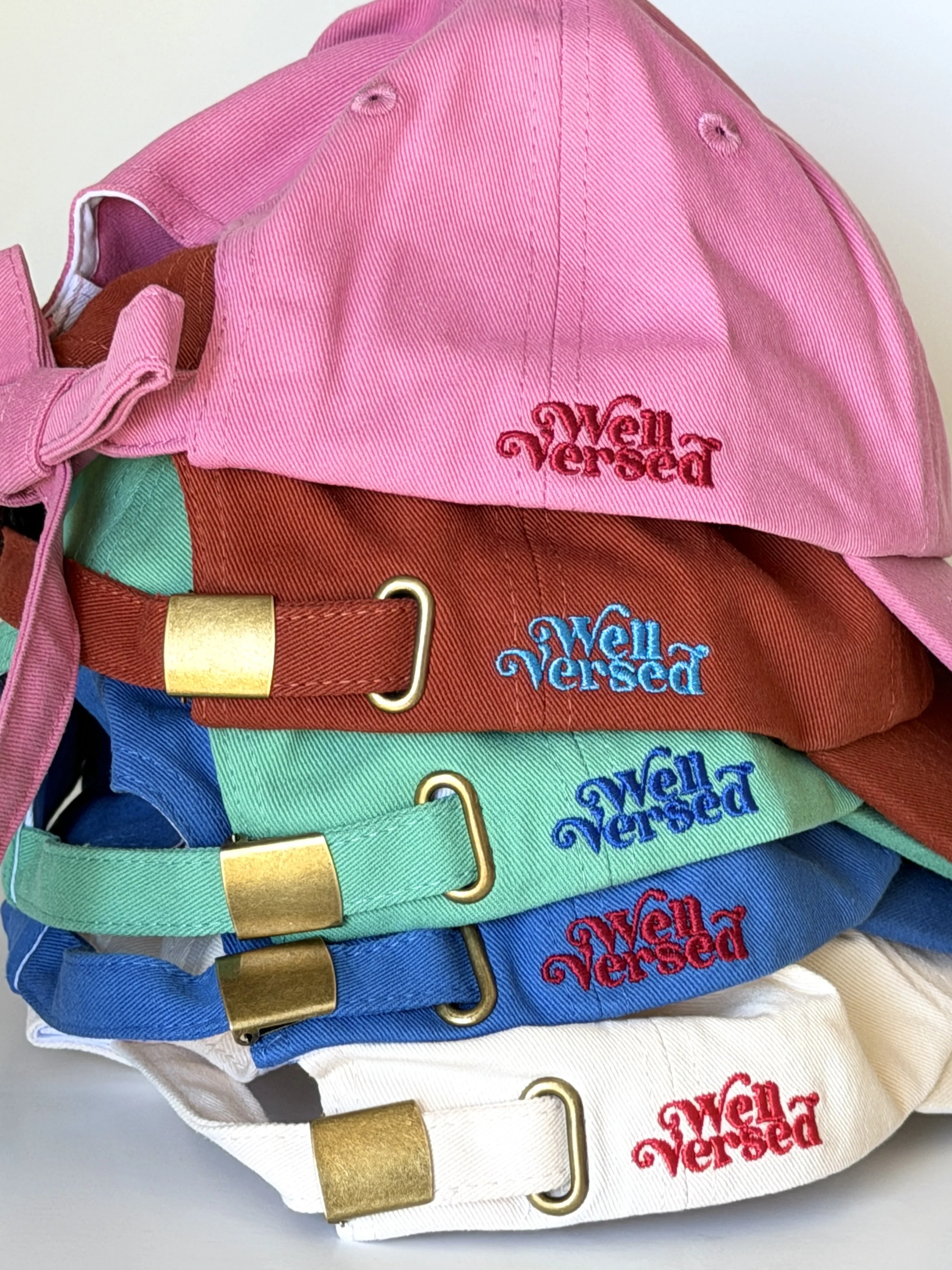 A stack of vibrantly colored hats with metal clasps and our brand logo Well Versed embroidered on the side