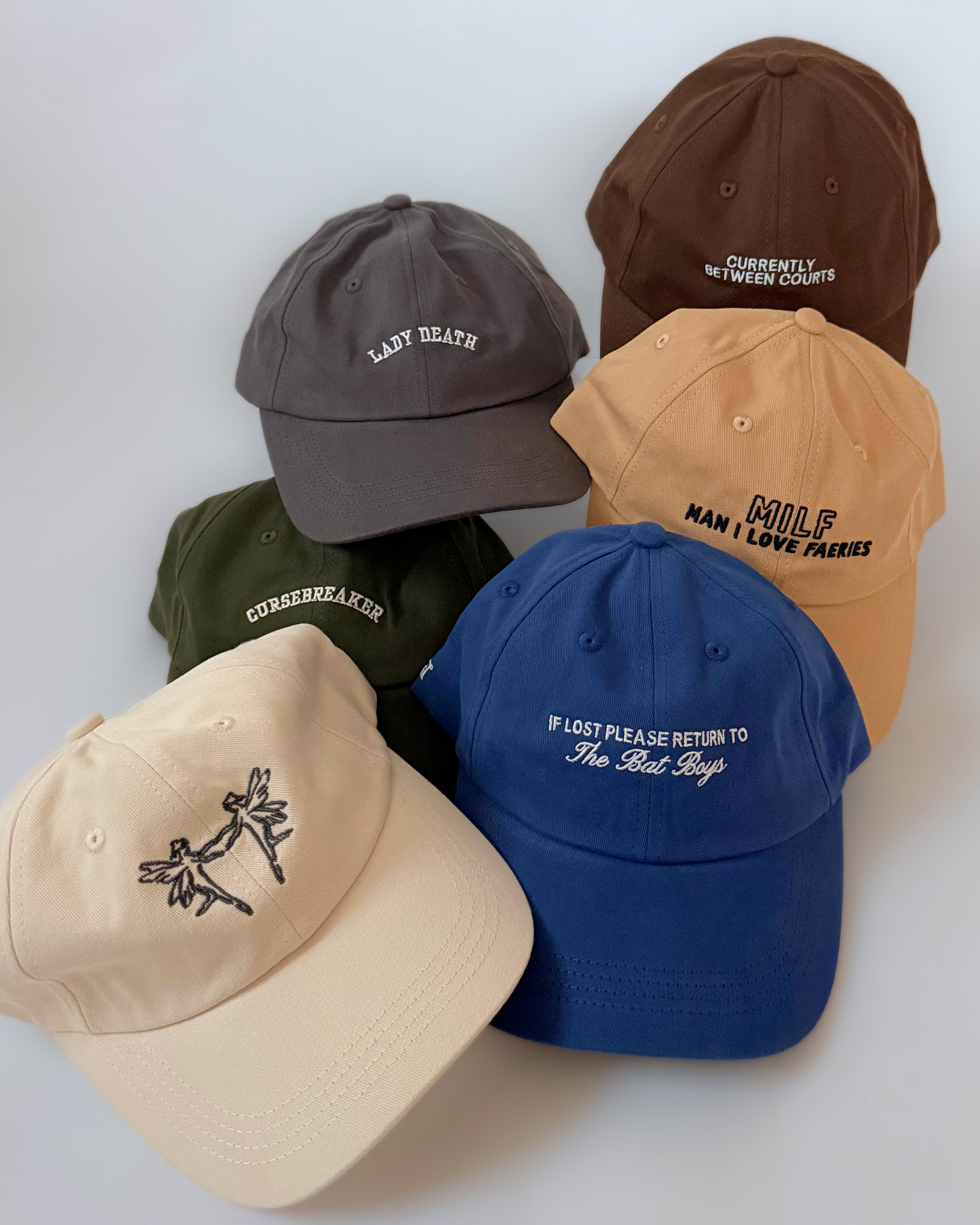 Two brown baseball caps showing cream “Currently Between Courts” embroidery on the front and embroidered stars beside an adjustable brass-tone clasp on the back.
