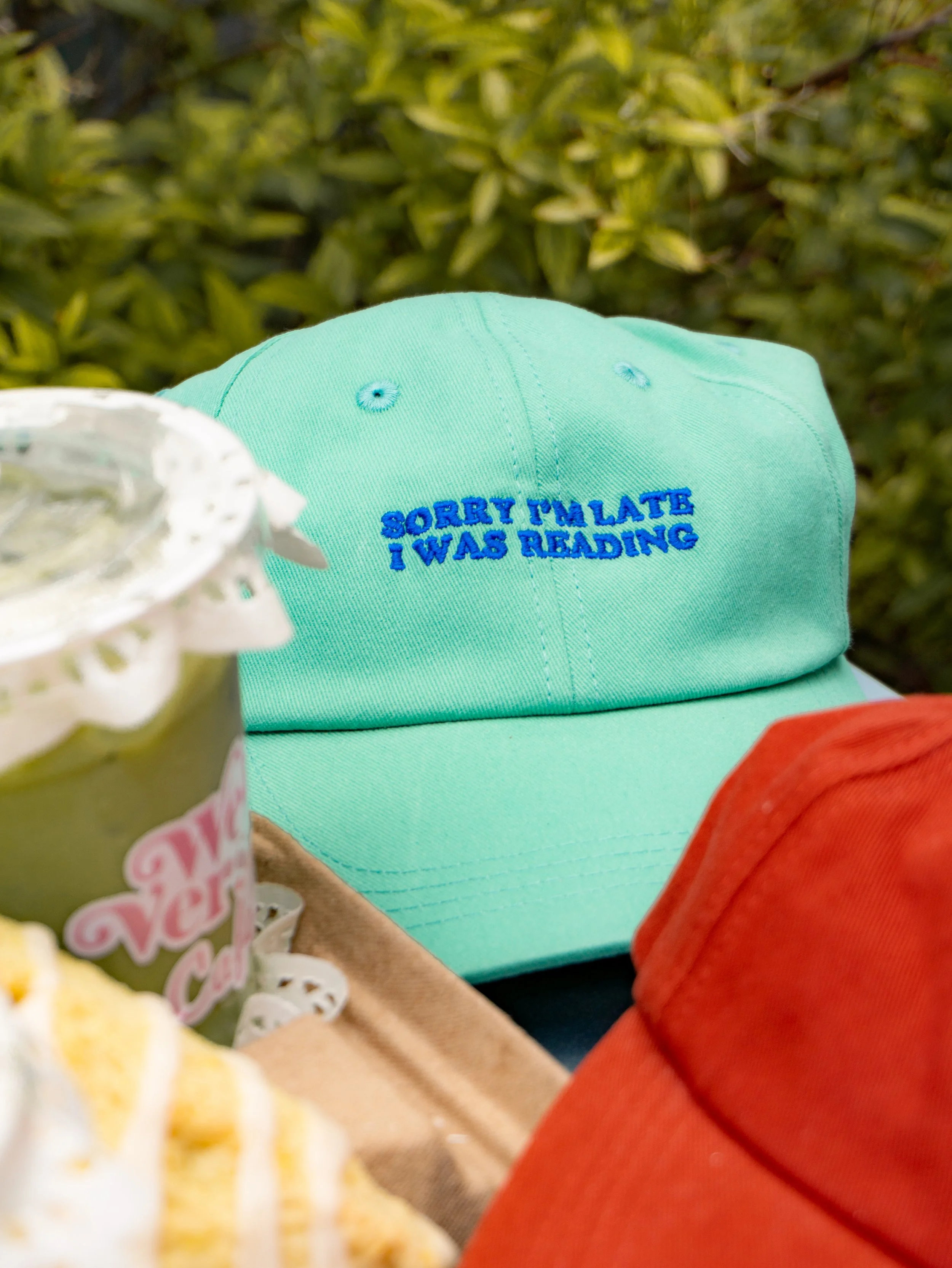 Mint green baseball cap embroidered with the phrase “SORRY I'M LATE I WAS READING” in bright blue lettering.