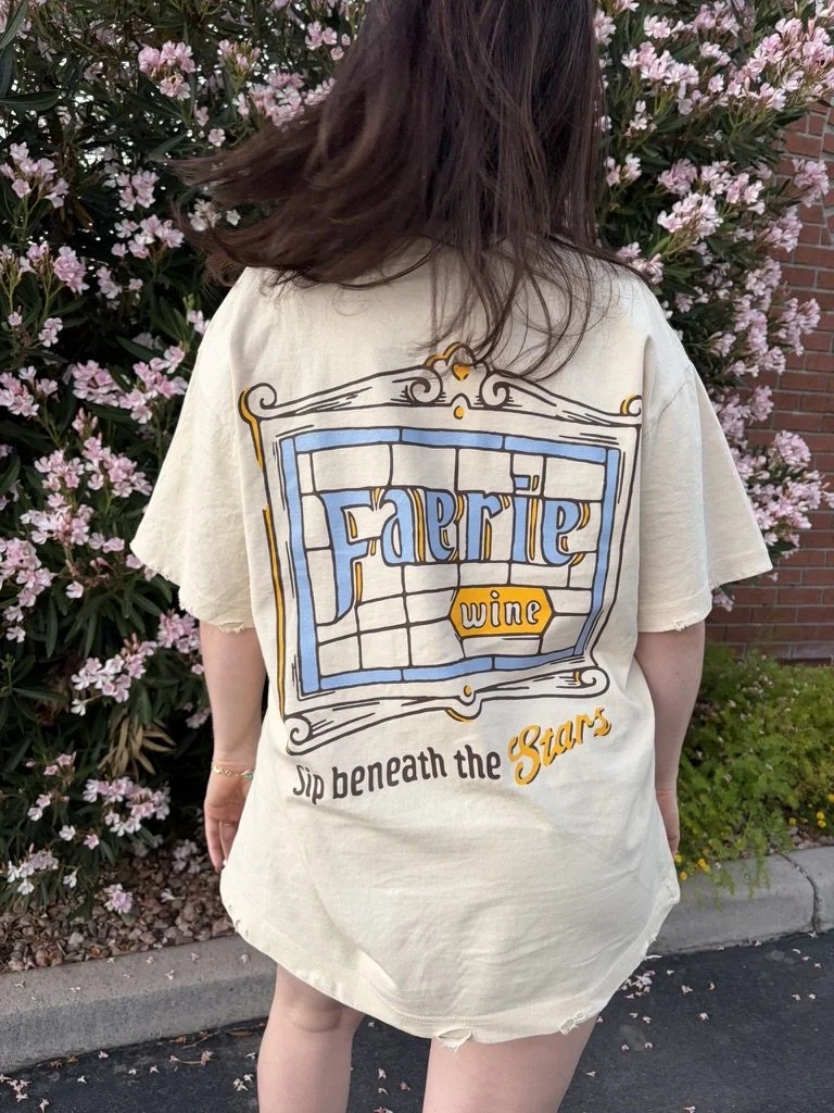 Back view of a model wearing an oversized ivory T-shirt with a blue, gold, and dark brown stained-glass-style graphic reading “Faerie Wine” and “Sip beneath the Stars."