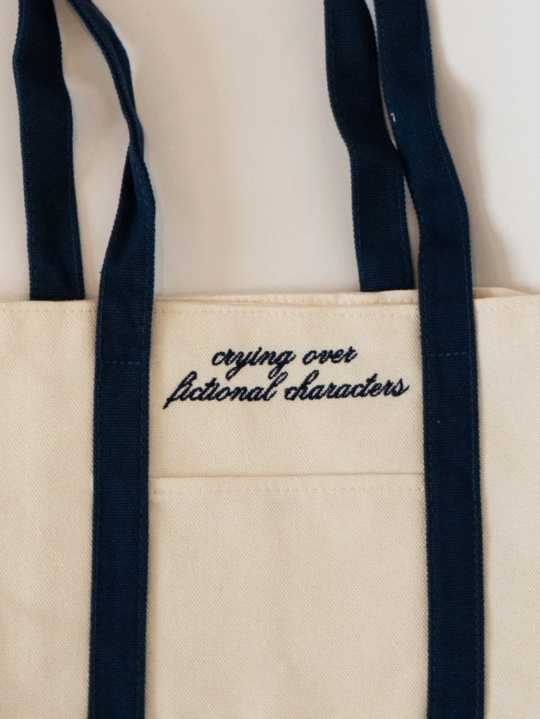 Crying Over Fictional Characters embroidered canvas tote bag with navy straps, a cute bookish tote for readers, romance book lovers, bookstore trips, and reader gifts.