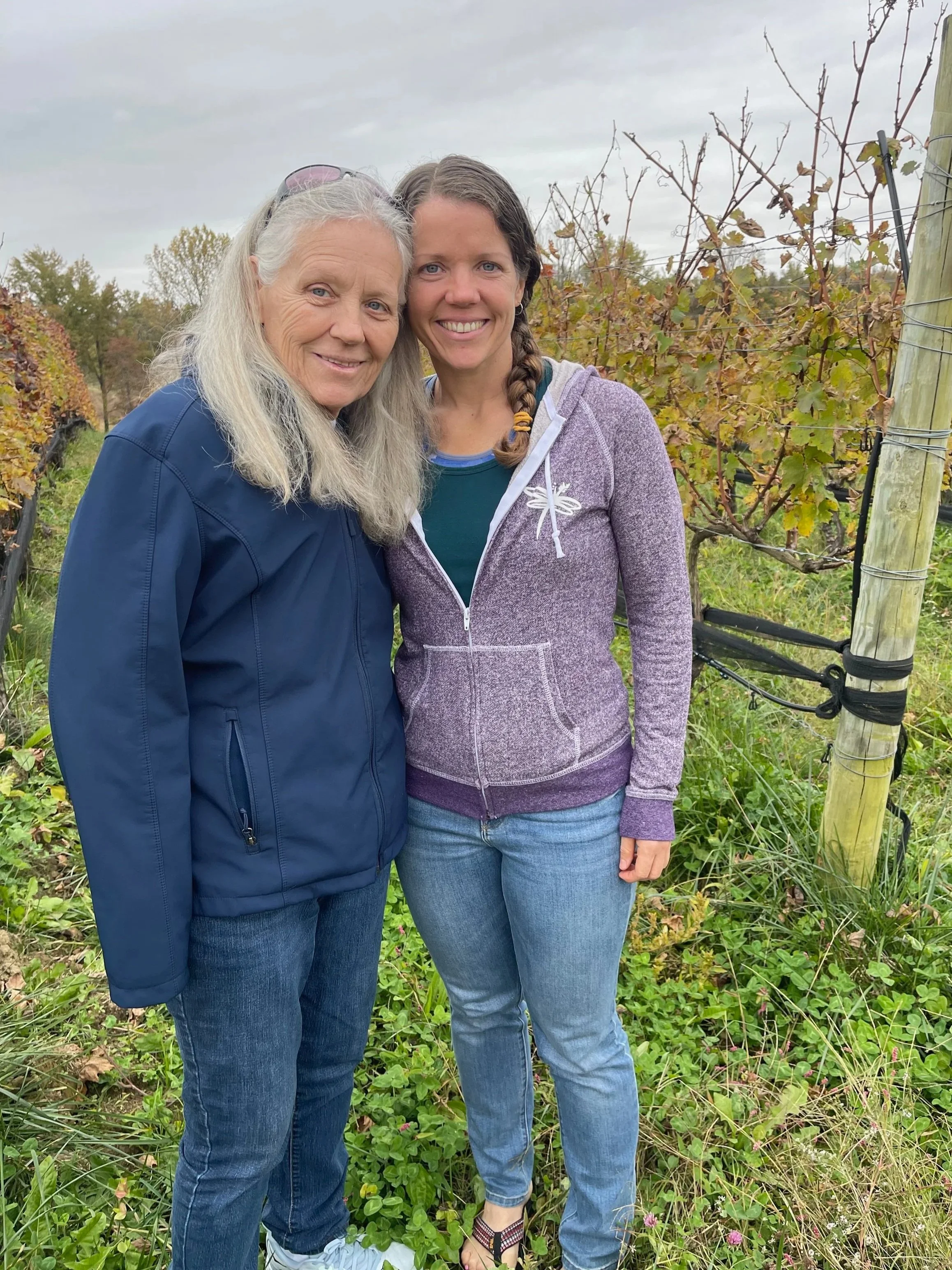An older white woman with long white and gray hair stands with her daughter, a white woman with long brown hair, in a vineyard.