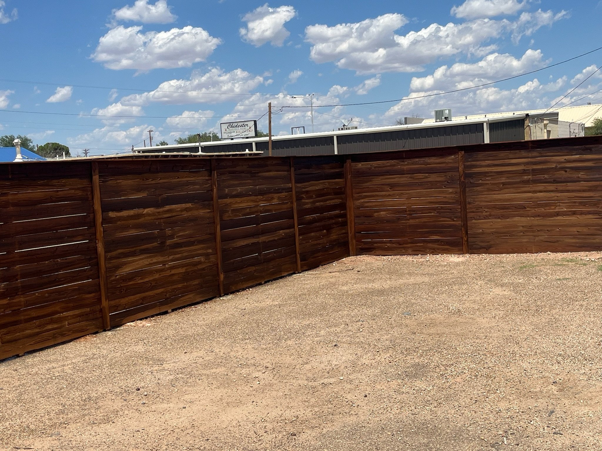 Wooden fence in a corner with gravel ground and cloudy blue sky.
