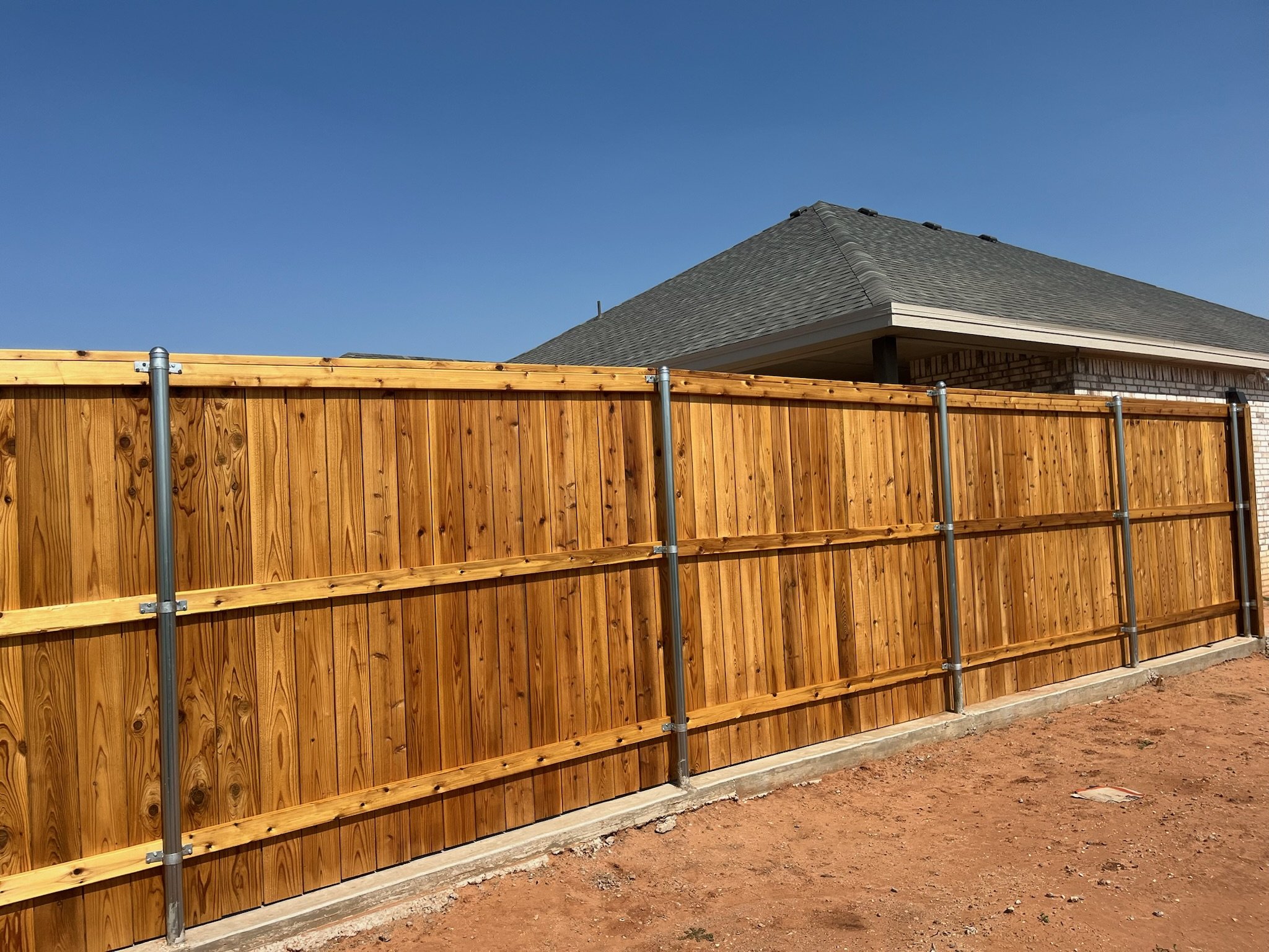 Wooden privacy fence with metal support posts, next to brick building under clear blue sky.