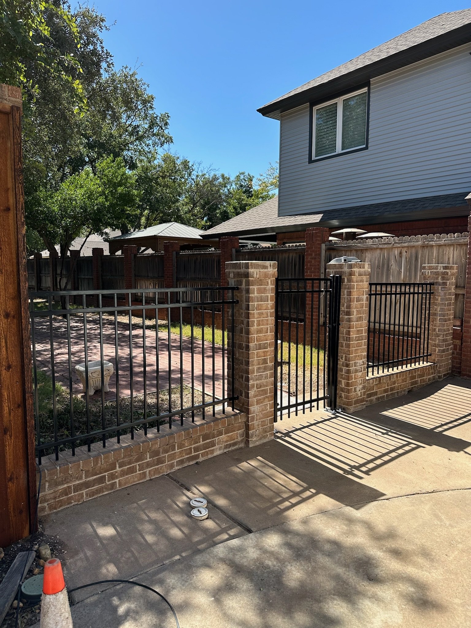 A residential area with a brick and black metal fence, including a gate. There are houses with pitched roofs and trees in the background, set under a clear blue sky.
