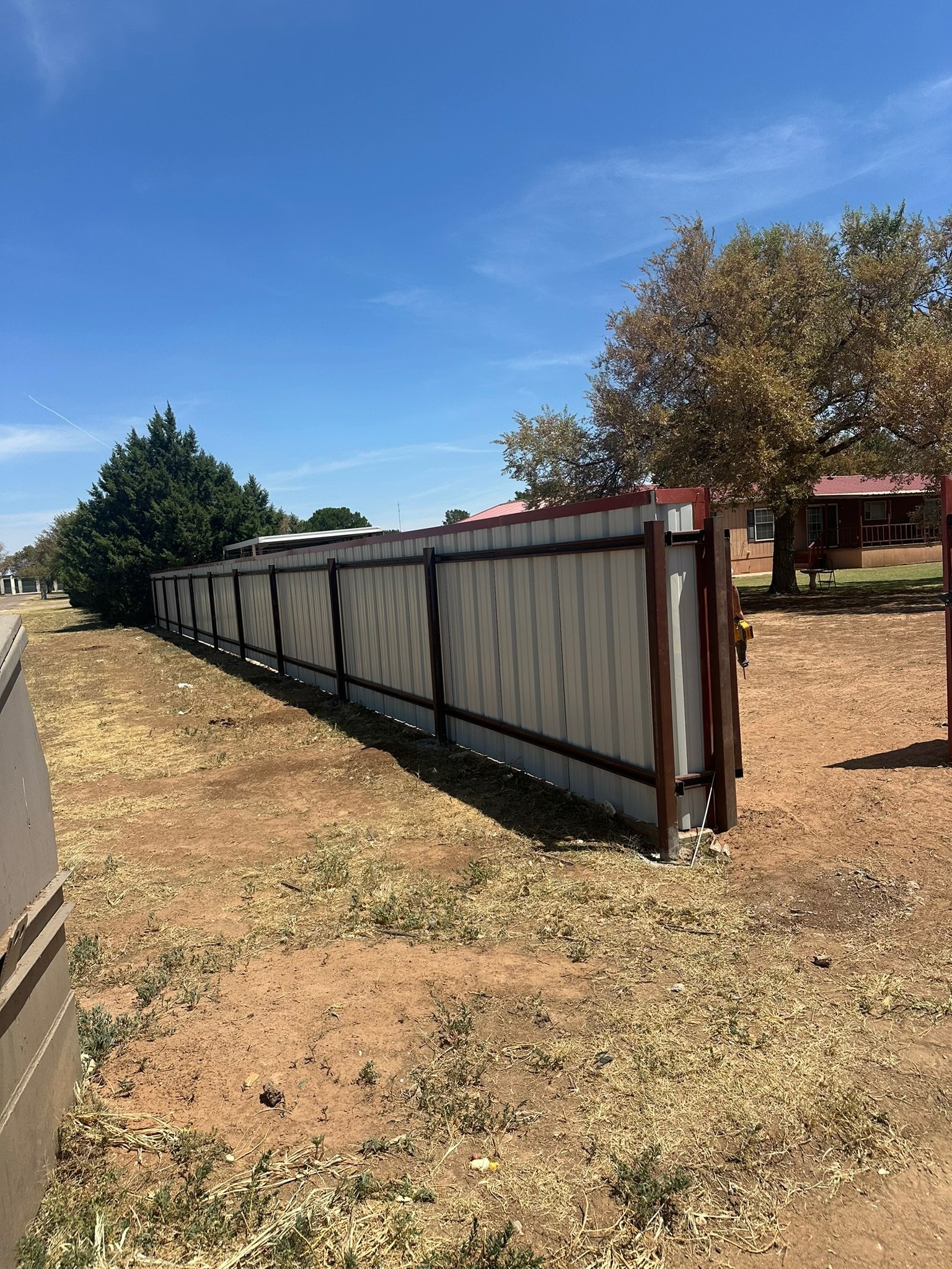 Metal fence on dry grass under a clear sky with trees and a house in the background.