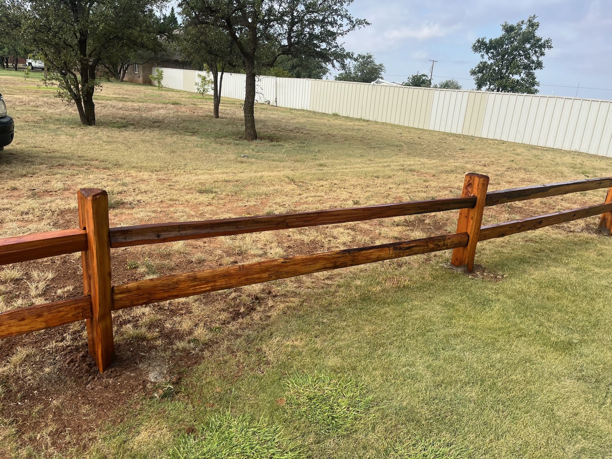 Wooden split rail fence along a grassy area with trees in the background and a white panel fence in the distance.
